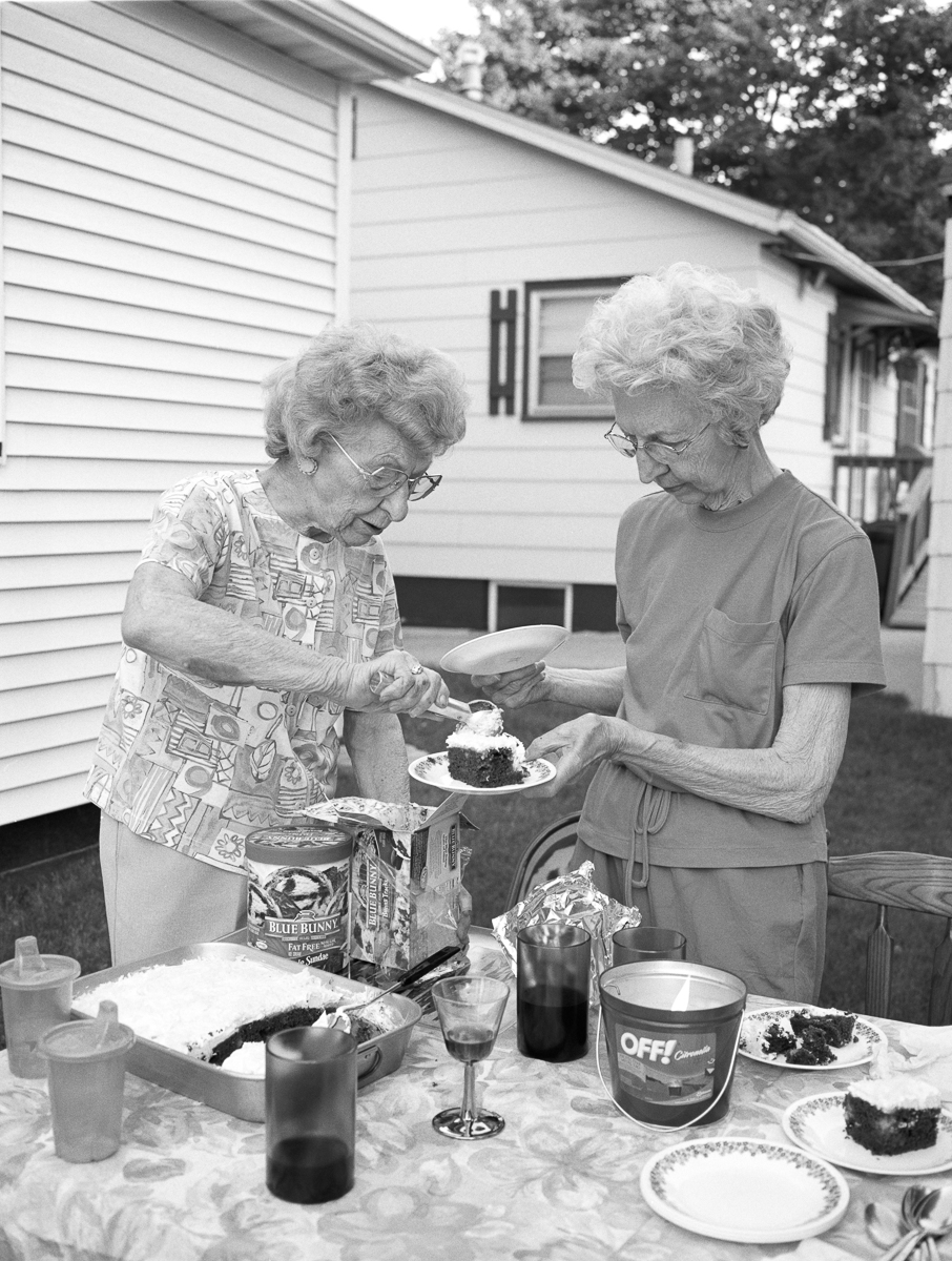 Two elderly women serve cake and ice cream at an outdoor table in front of a house, captured in black and white. The scene shows a casual gathering with various food items, beverages, and tableware arranged on a patterned tablecloth.