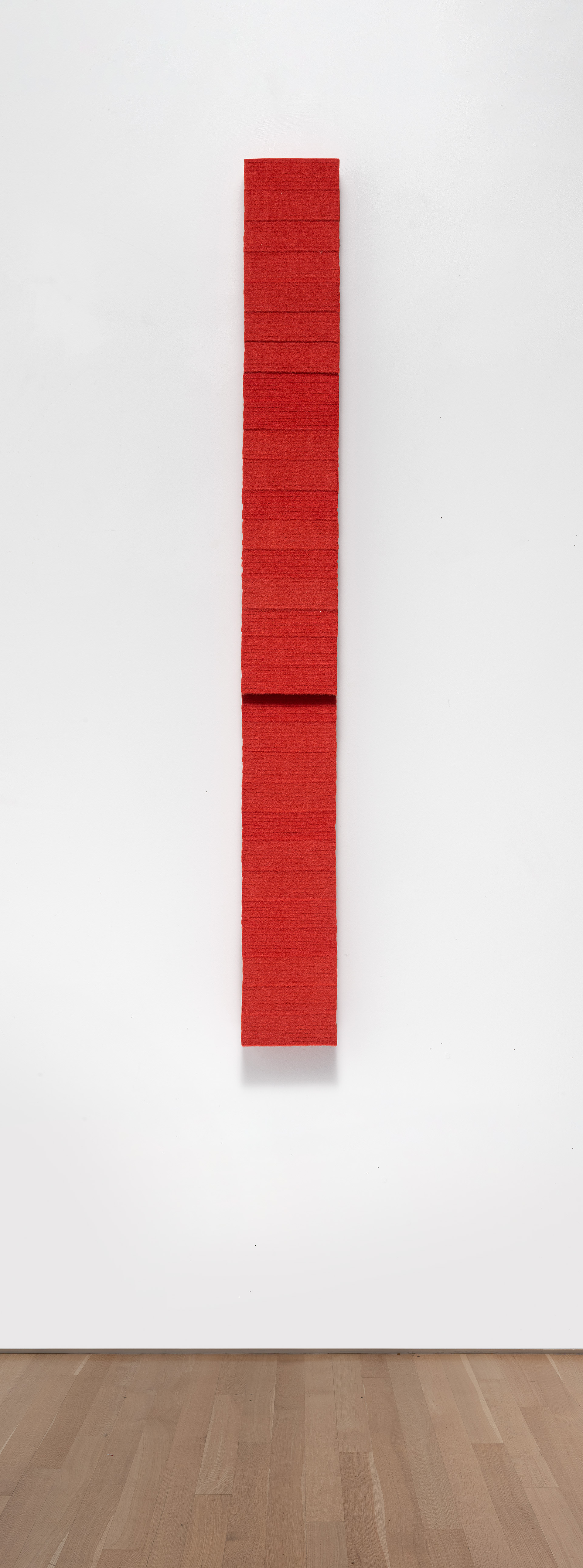 A vertical red wool felt rectangle mounted on a white gallery wall, composed of stacked rectangular segments with subtle horizontal divisions creating a modular, column-like structure.