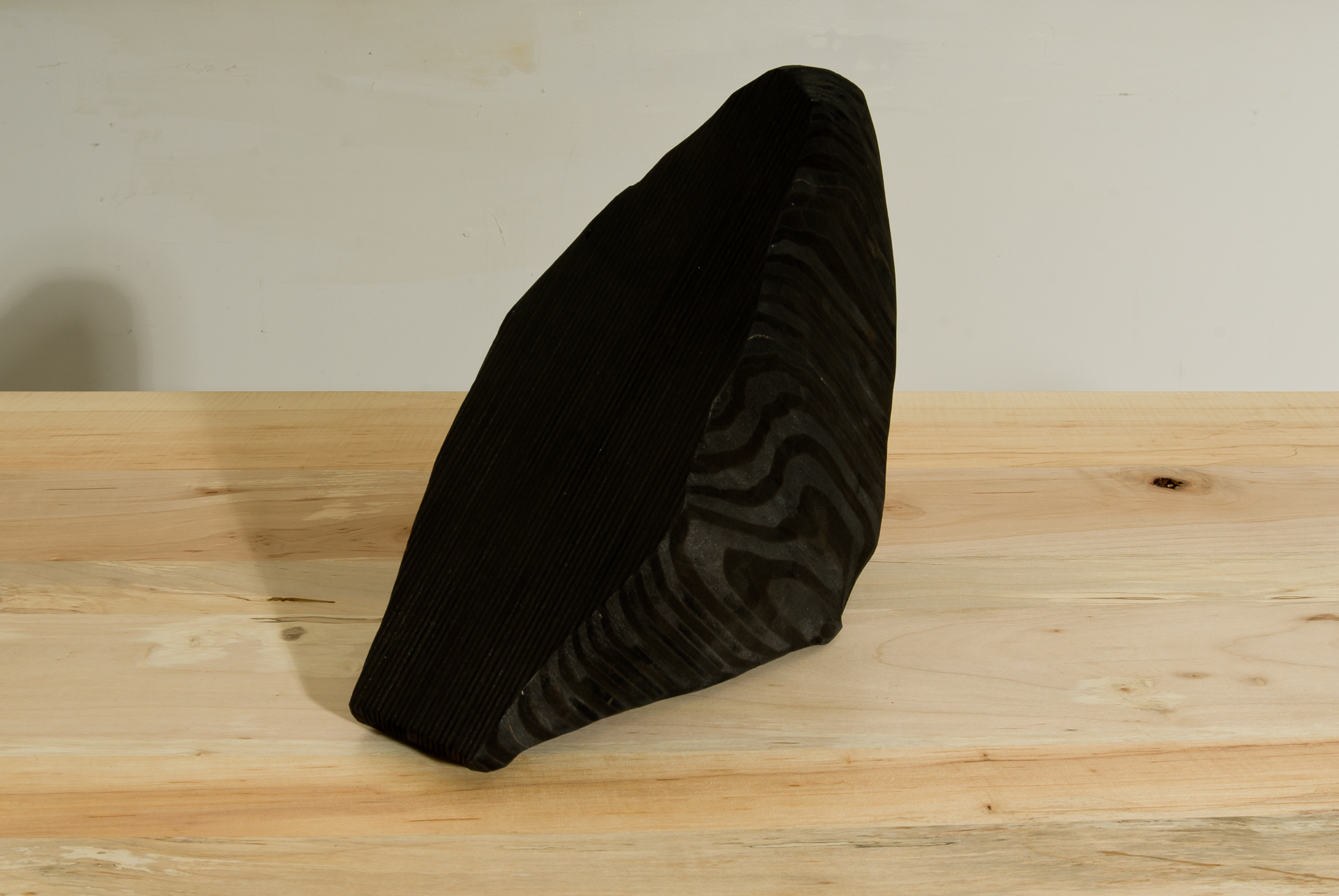 A black stained wooden sculpture tilted at an angle on a light wooden floor against a white wall. The form is organic and curved, resembling a natural shell or fossil, with subtle ridged texturing across its surface.