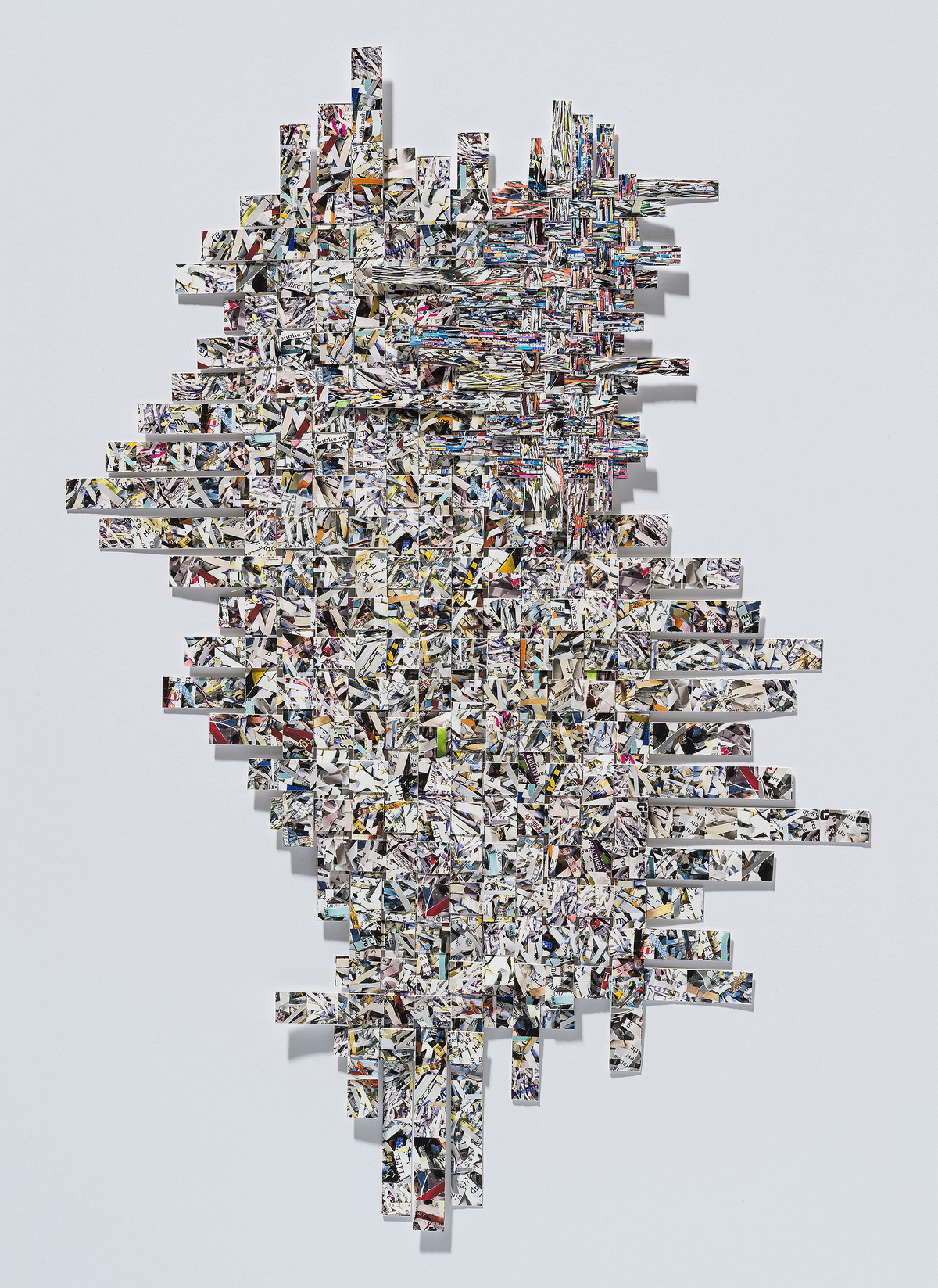 A large-scale collage composed of hundreds of rectangular strips of printed images arranged in a staggered, woven pattern against a white wall. The strips, predominantly black and white with occasional color accents, create a dense, interlocking composition that shifts between structured geometric formations and organic, flowing arrangements. Shadows cast by the protruding elements add depth and dimensionality to the work.