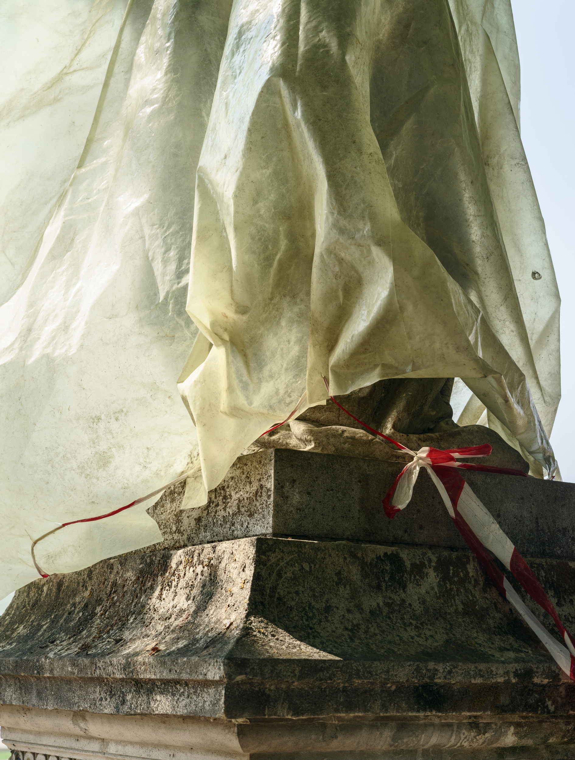 Pale fabric drapes over a weathered stone base, with a red and white ribbon tied around the lower section. The translucent material catches light, creating soft shadows and folds against the textured, lichen-covered stone beneath a bright sky.