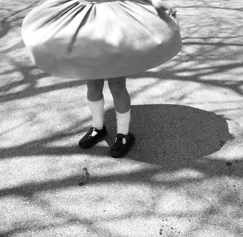 Black and white photograph of a child's lower body twirling in a full skirt on a concrete surface, casting a distinct shadow. The child wears white socks and dark Mary Jane shoes, captured mid-motion with the skirt billowing outward.