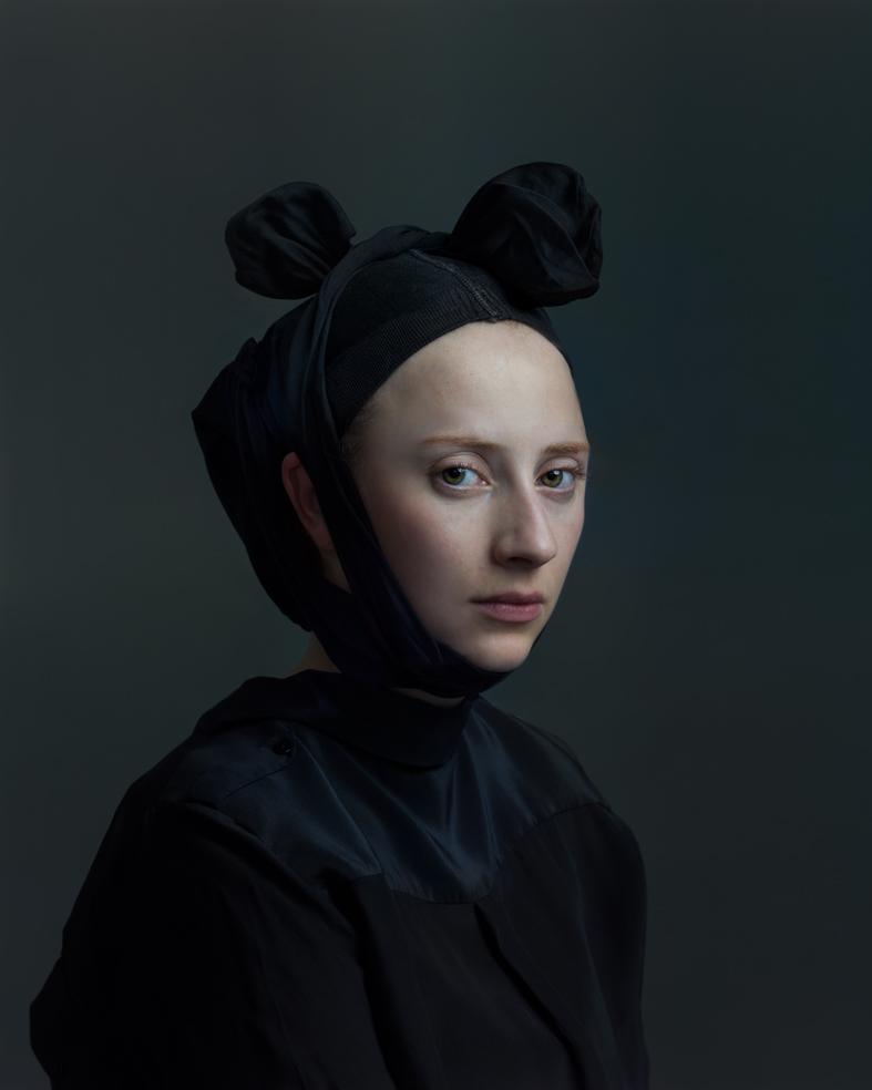 Portrait of a person with a pale complexion facing slightly left against a muted teal-gray background, wearing all black clothing and a black headwrap styled with two sculptural fabric buns at the crown. The lighting is soft and directional, emphasizing the contours of the face and creating a contemplative mood.
