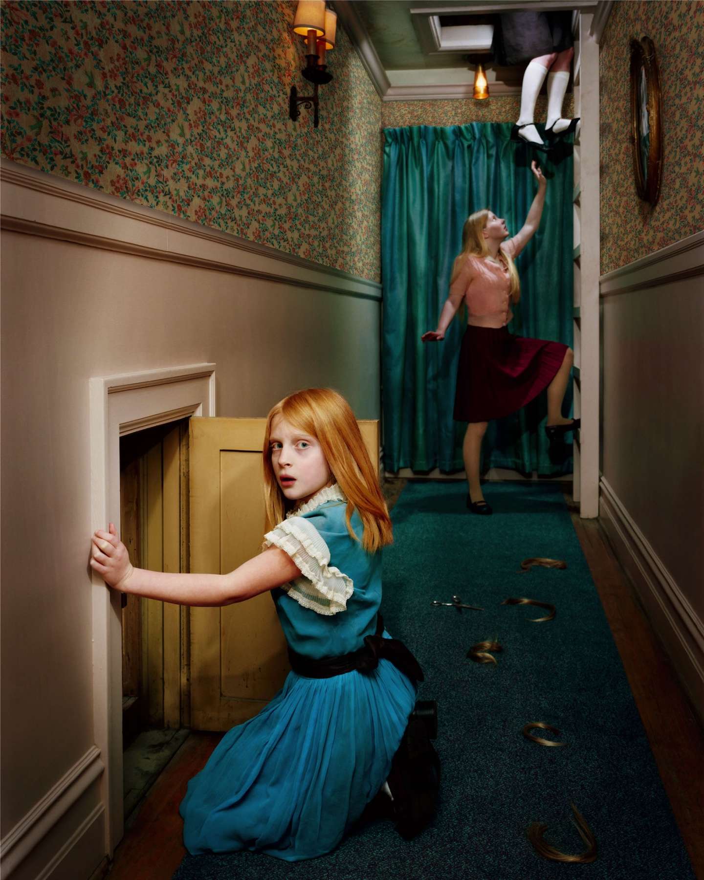 Two red-haired figures in a narrow hallway with floral wallpaper and teal carpet. In the foreground, a young girl in a blue dress with white ruffled collar kneels by a yellow door, looking back toward the camera. In the background, a shirtless figure reaches upward toward hanging white objects near teal curtains. Warm sconces and a ceiling fixture illuminate the scene, creating an eerie, dreamlike domestic interior.