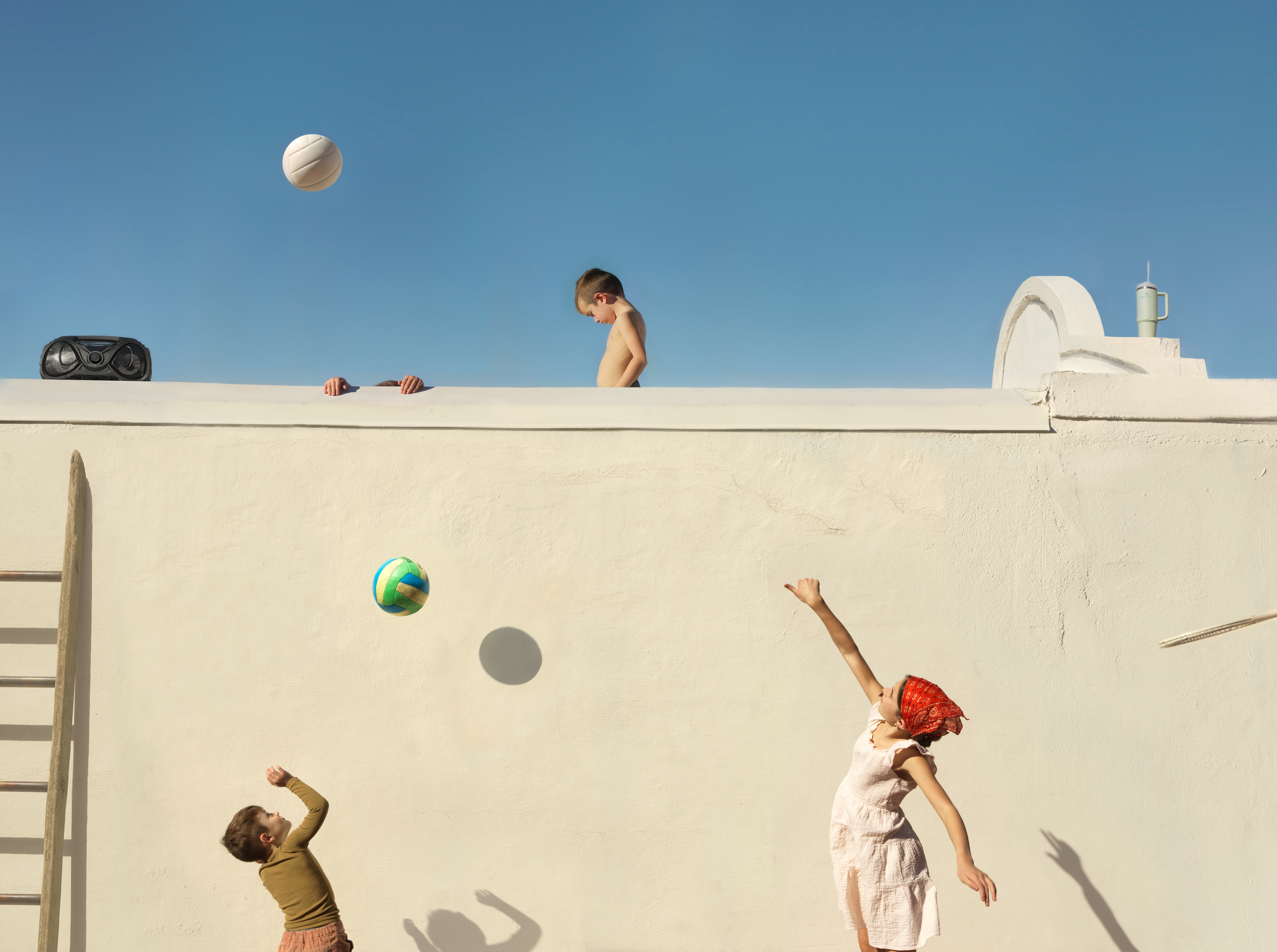 Children play with balls on a stark white concrete wall dividing an upper and lower level against a clear blue sky. A shirtless boy stands on top of the wall watching, while below, two children toss colorful balls upward, their shadows cast sharply against the pale surface. Beach equipment and a white structure are visible at the top edge of the composition.