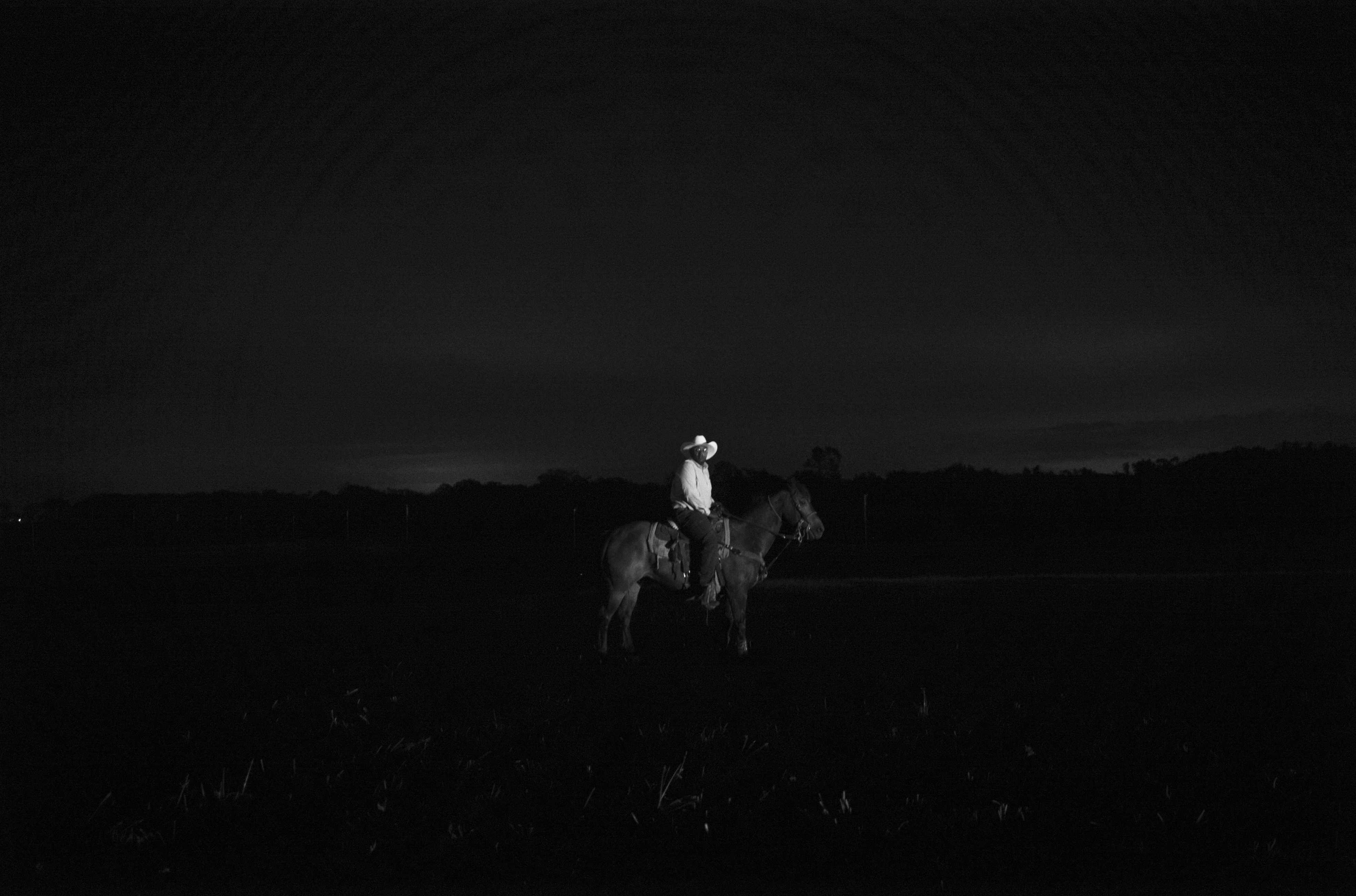 A solitary figure in light clothing sits atop a horse in an expansive, dark landscape at dusk or night. The composition is rendered in high-contrast black and white, with the rider and horse illuminated against a nearly black sky and terrain, creating a stark, isolated presence on the plains.