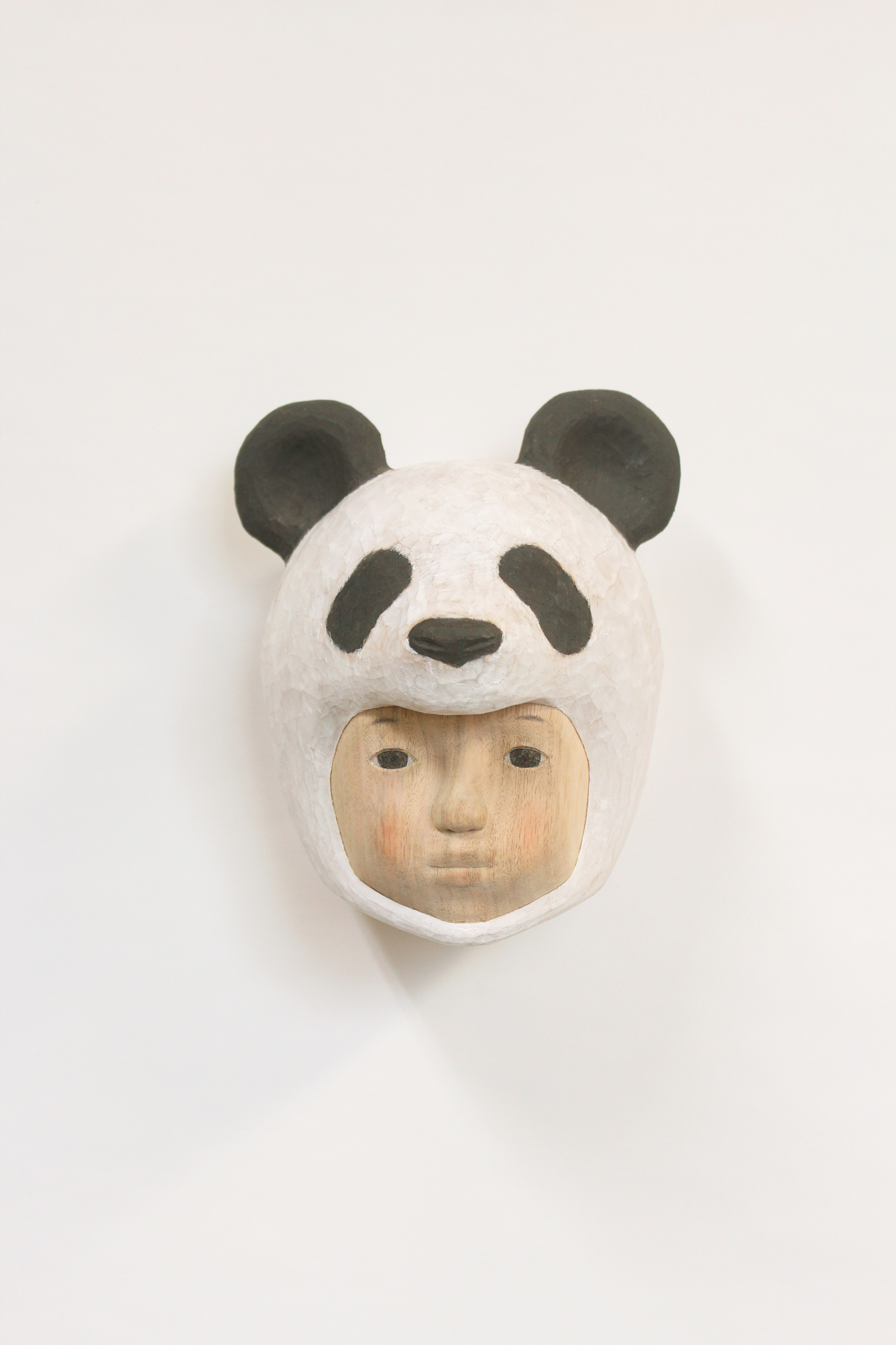A carved wooden mask depicting a human face wearing a giant panda head covering, with black and white coloring and subtle flesh-toned facial features below. The sculpture combines naturalistic human portraiture with stylized animal characteristics, mounted on a white background.