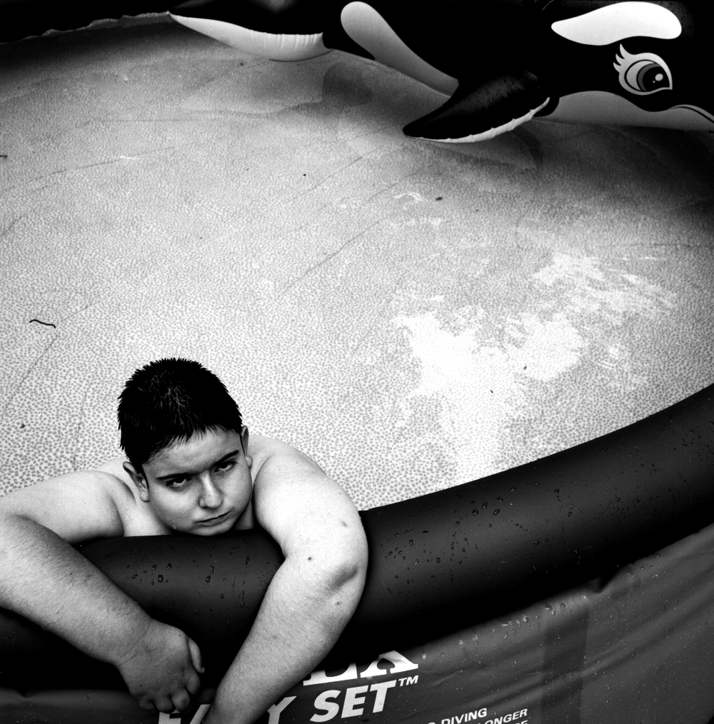 Black and white photograph of a young boy resting against an inflatable pool toy shaped like a killer whale, looking downward with a contemplative expression. The composition captures the boy partially submerged in shallow water with textured pool surface visible in the background.