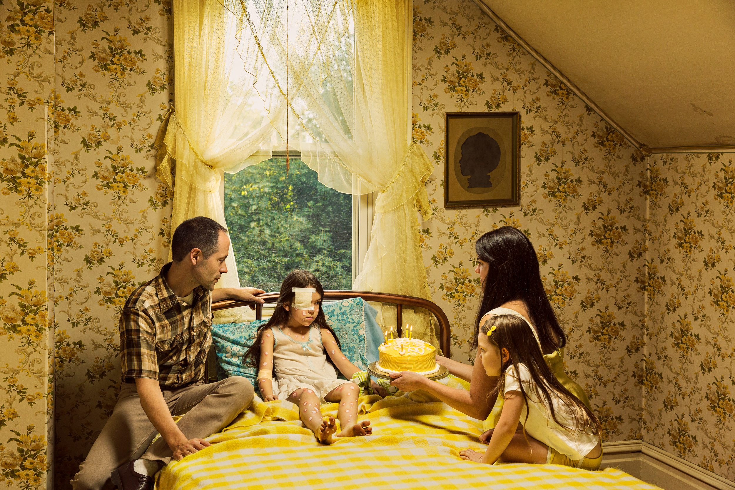 A family of four sits on a yellow striped bed in an attic room with floral wallpaper and yellow curtains framing a window overlooking green foliage. A yellow birthday cake with lit candles sits on the bed as the father touches his young daughter's head while the mother and older daughter look on.