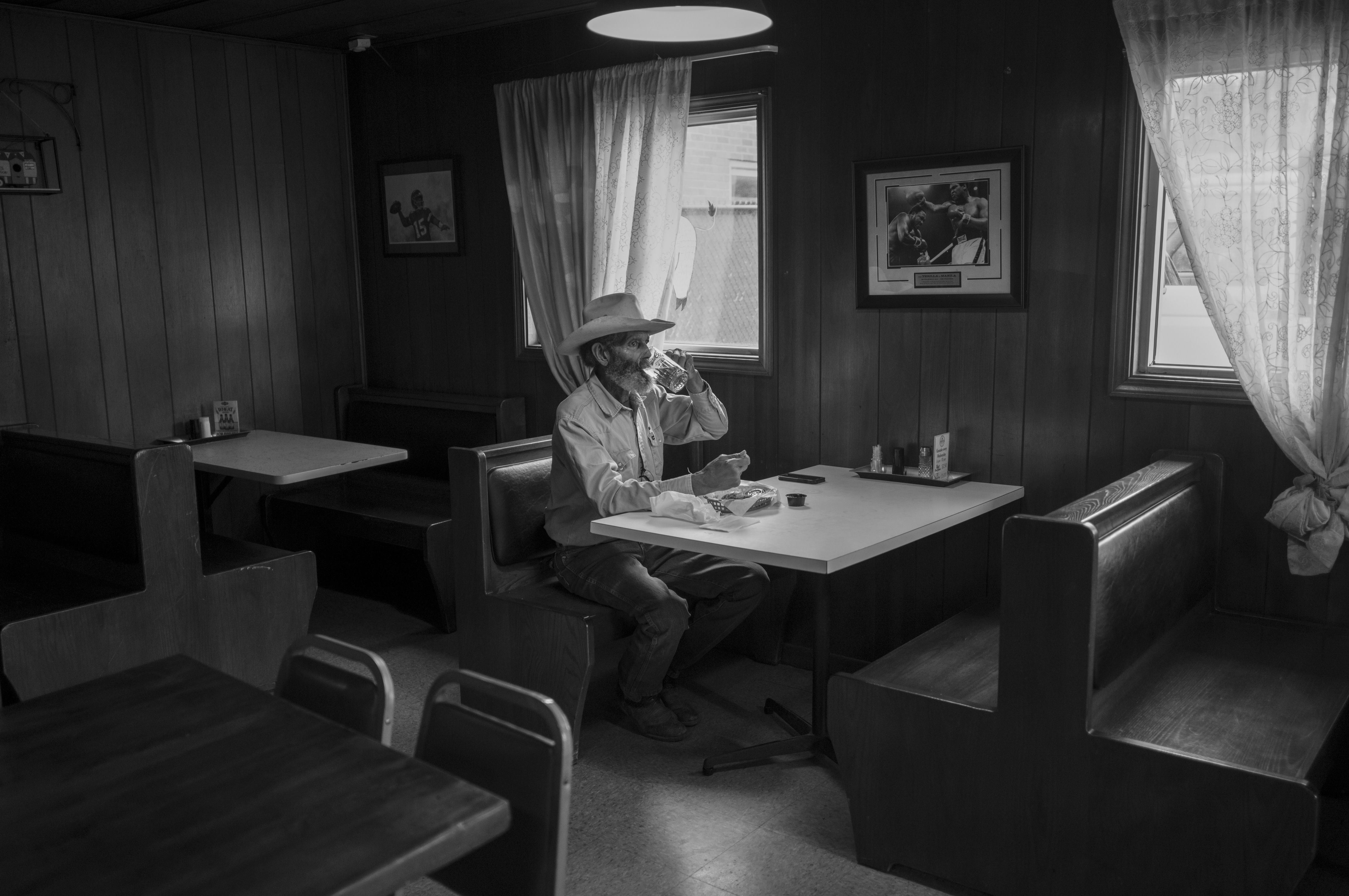 Black and white photograph of a solitary man wearing a cowboy hat seated at a booth table in a modest diner interior, drinking from a cup. The dimly lit space features vinyl booth seating, framed photographs on wood-paneled walls, sheer curtains filtering light through windows, and a recessed ceiling light, creating a stark, contemplative atmosphere.