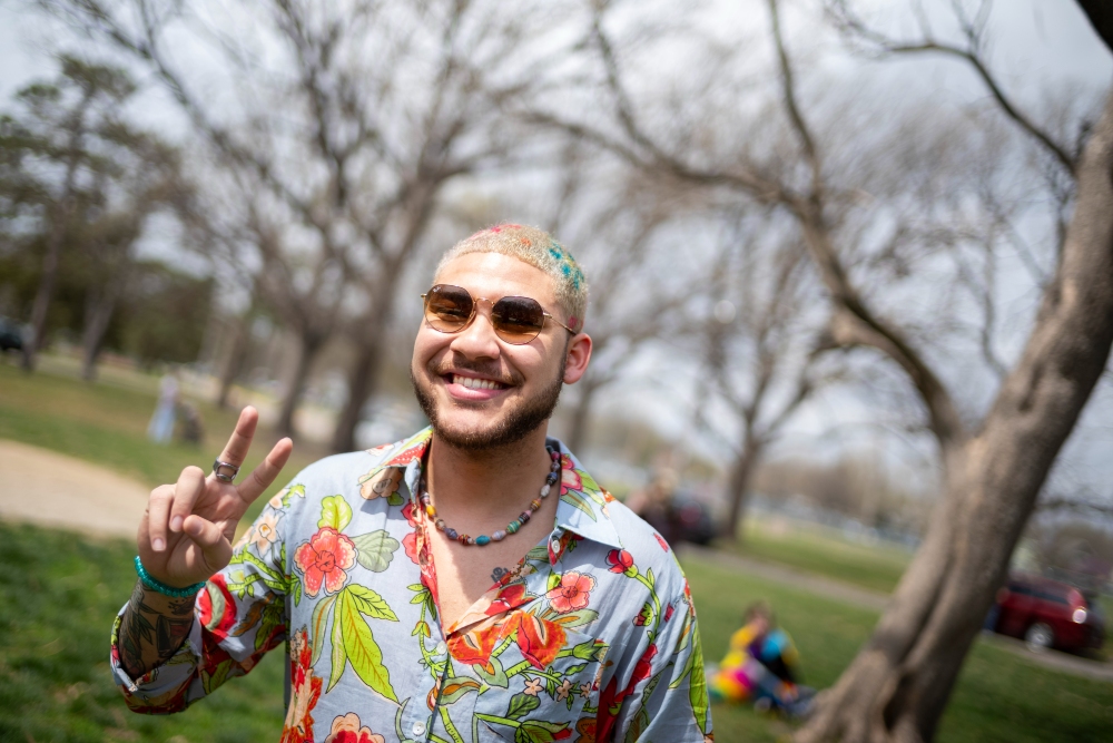 A person wearing sunglasses and a colorful floral shirt smiles at the camera while making a peace sign, standing in a park with bare trees in the background.