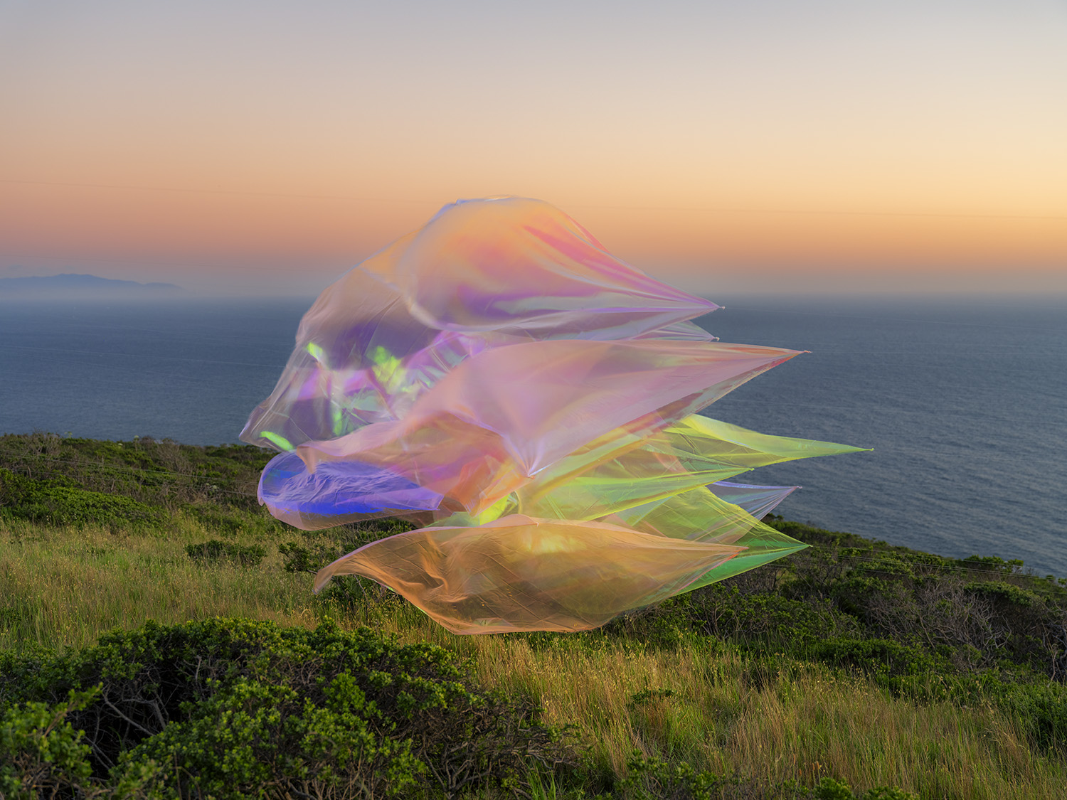 Iridescent, translucent organza fabric billows across a coastal hillside at sunset, its layered folds catching light in shimmering gradients of pink, purple, yellow, and green. The luminous sculptural form floats above wild grasses and shrubs with the ocean and twilight sky visible in the background.