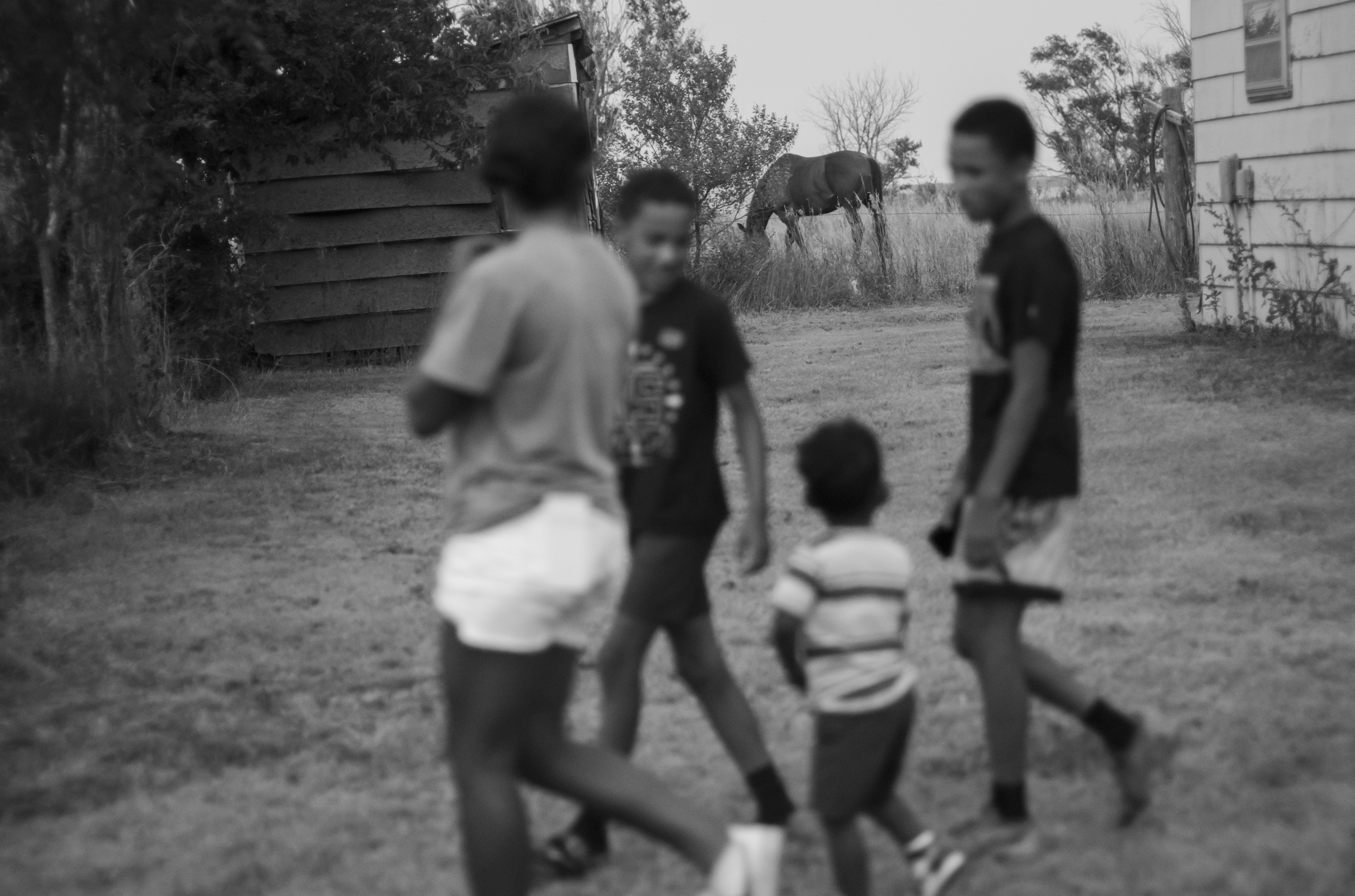 Black and white photograph of several figures in motion in a rural dirt yard between weathered wooden structures and overgrown vegetation. The subjects are intentionally blurred, creating a sense of movement and ethereal quality, while the architectural elements and landscape remain in sharper focus.