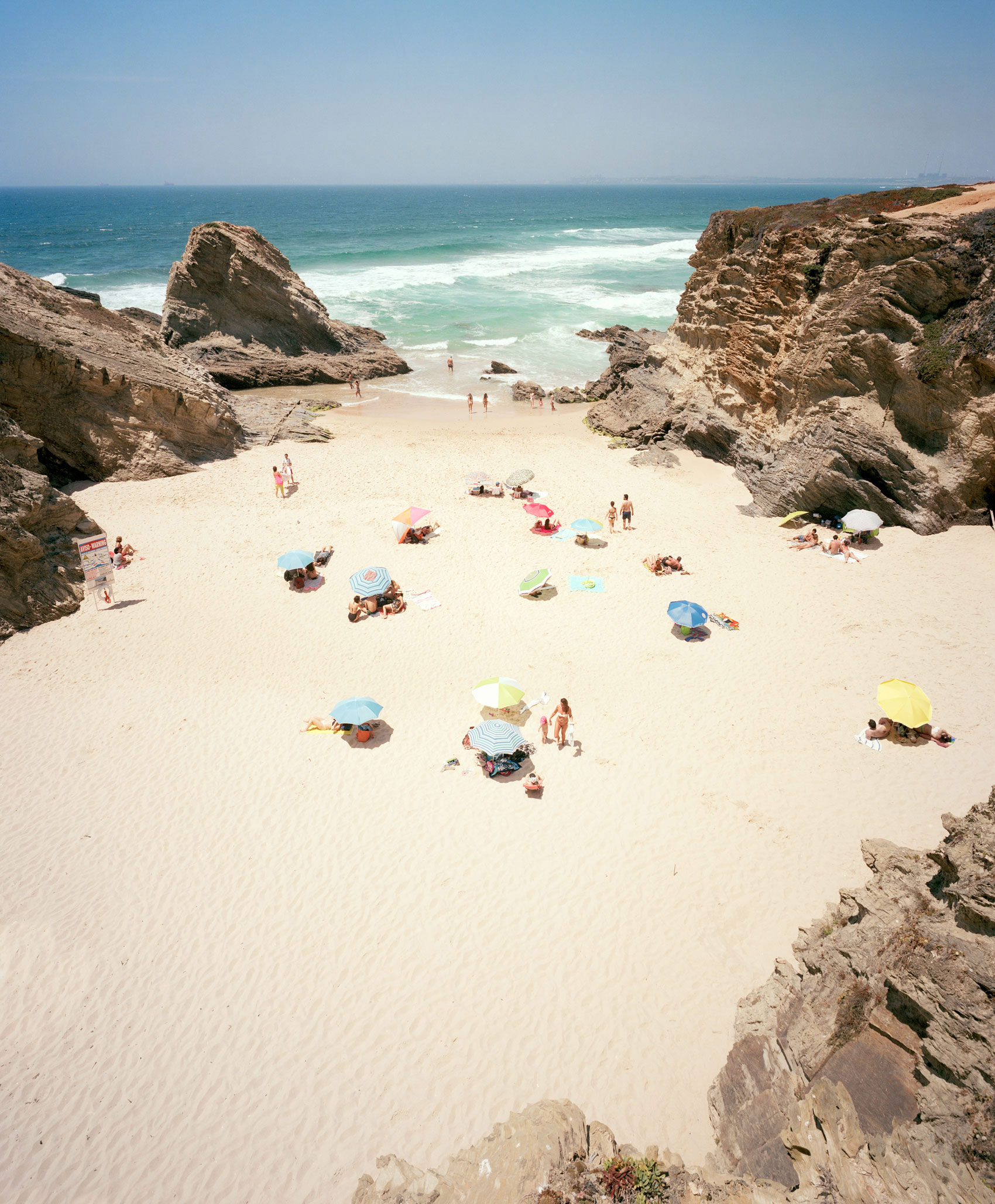 Aerial view of a secluded sandy beach nestled between towering limestone cliffs, with scattered beachgoers sheltering beneath colorful umbrellas in shades of blue, yellow, pink, and green. Turquoise waves break along the shoreline under a clear sky, creating a serene composition that captures the scale of the natural landscape against small human figures below.