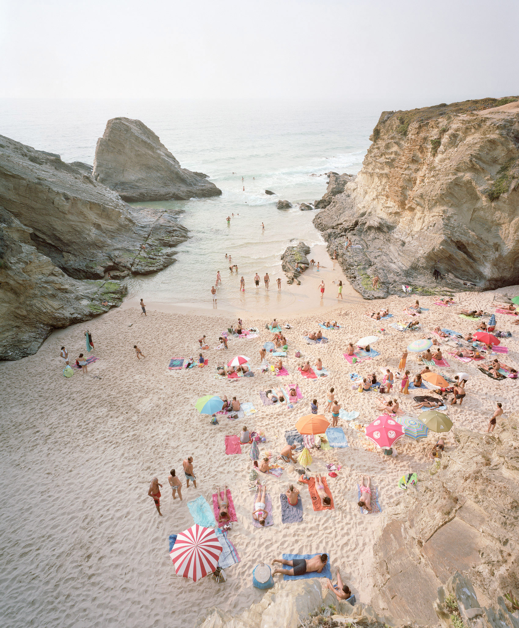 Aerial view of a crowded beach nestled between towering chalk cliffs, with numerous beachgoers scattered across the sand beneath colorful umbrellas and towels in shades of pink, red, blue, and yellow, while others wade in the shallow turquoise waters.