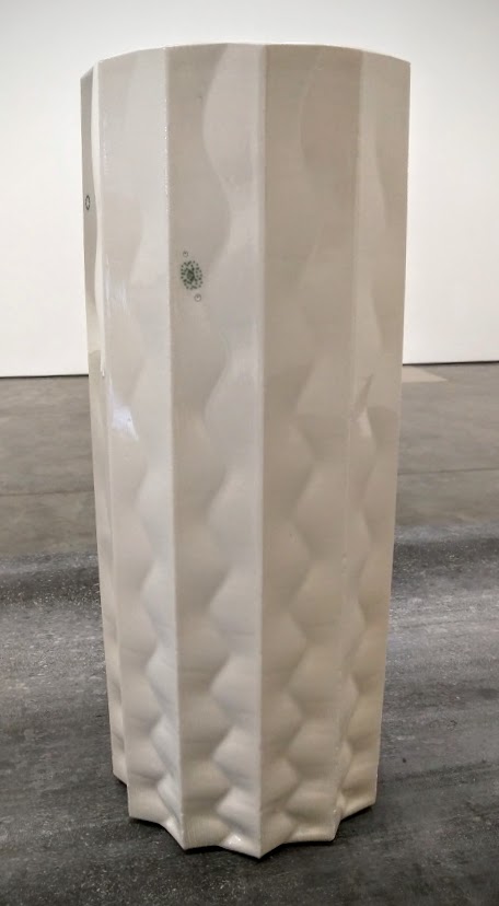 A tall, cylindrical white porcelain vessel with undulating vertical ridges and a small green circular mark near the top, displayed against a neutral gallery floor and wall.