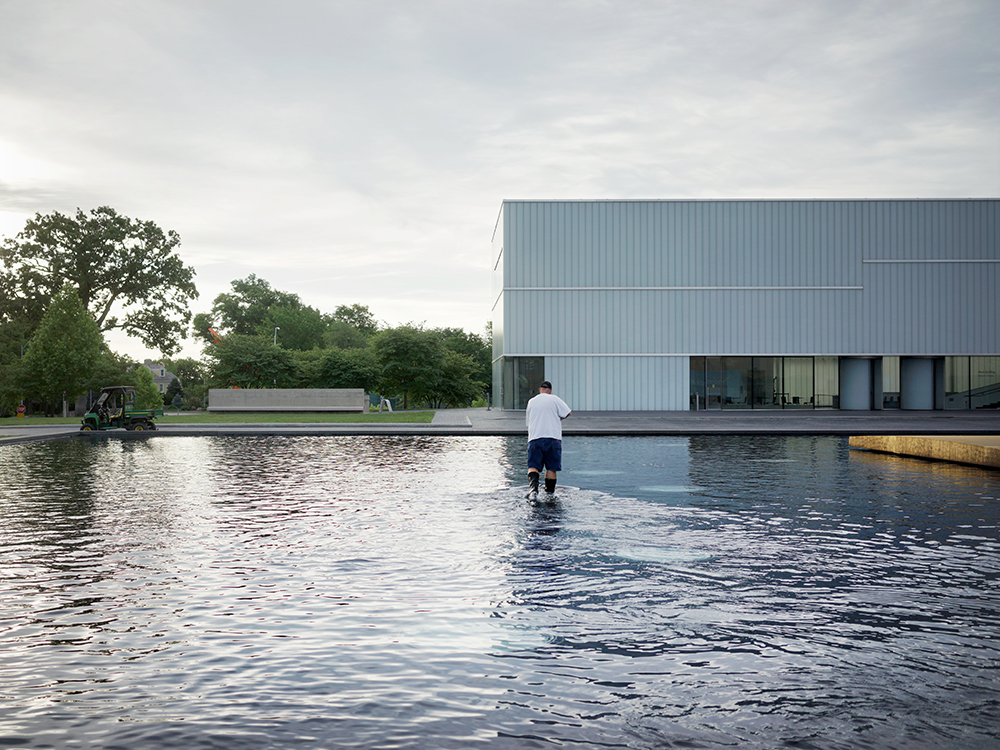 A person in white shirt and dark pants wades through shallow water toward a modernist building with corrugated metal cladding and horizontal windows. Trees line the far bank of the water, while industrial equipment operates on the left side under overcast sky.