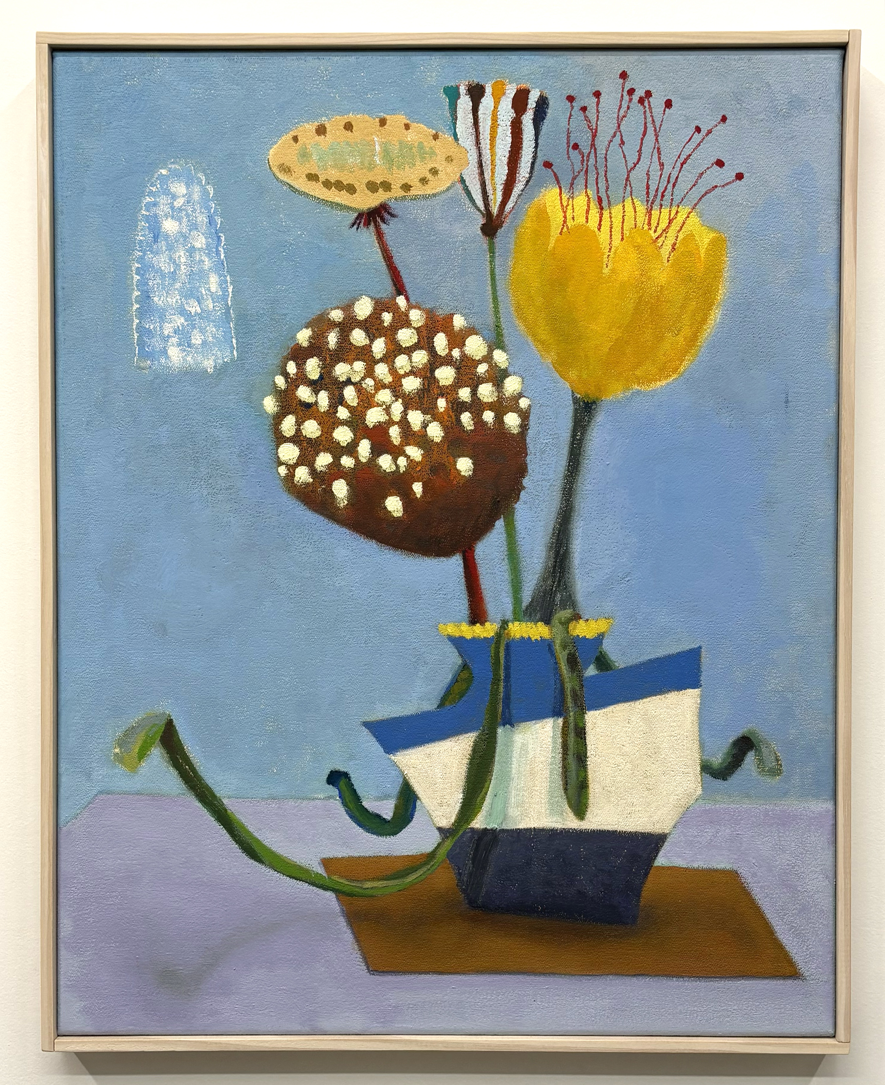 A still life painting depicting three stylized flowers with distinctive blooms—a pale yellow oval form with dark spots, a dark brown sphere dotted with cream-colored circles, and a bright yellow rounded shape with thin red stamens—arranged in a decorative blue and white geometric vase against a soft blue background. A withered stem curves from the base, and a faint white form appears in the upper left corner.
