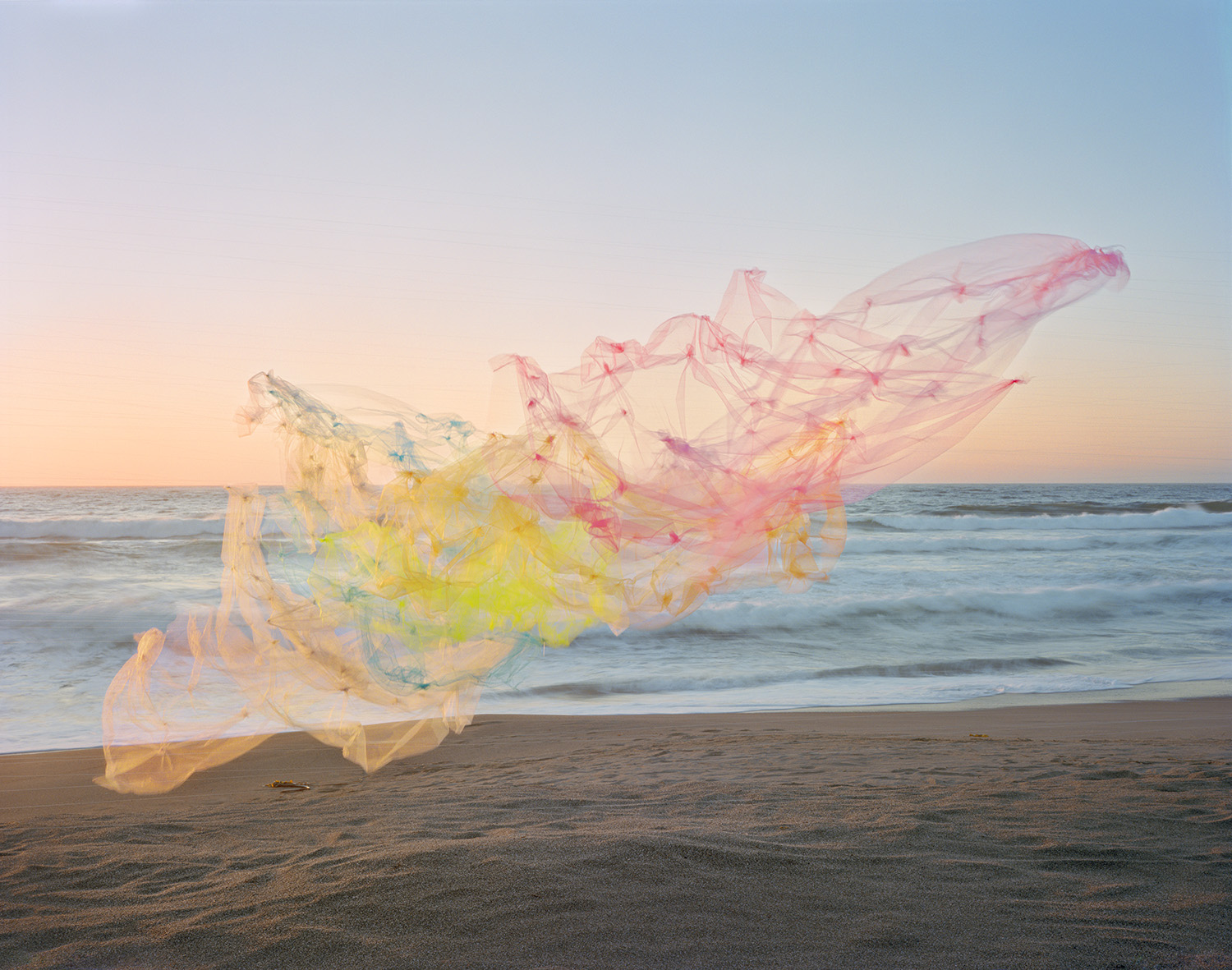 Billowing tulle fabric in soft gradations of pink, yellow, and blue floats across a beach at golden hour, with the ocean and horizon visible beyond. The translucent, flowing material creates an ethereal, sculptural form against the sand and seascape.