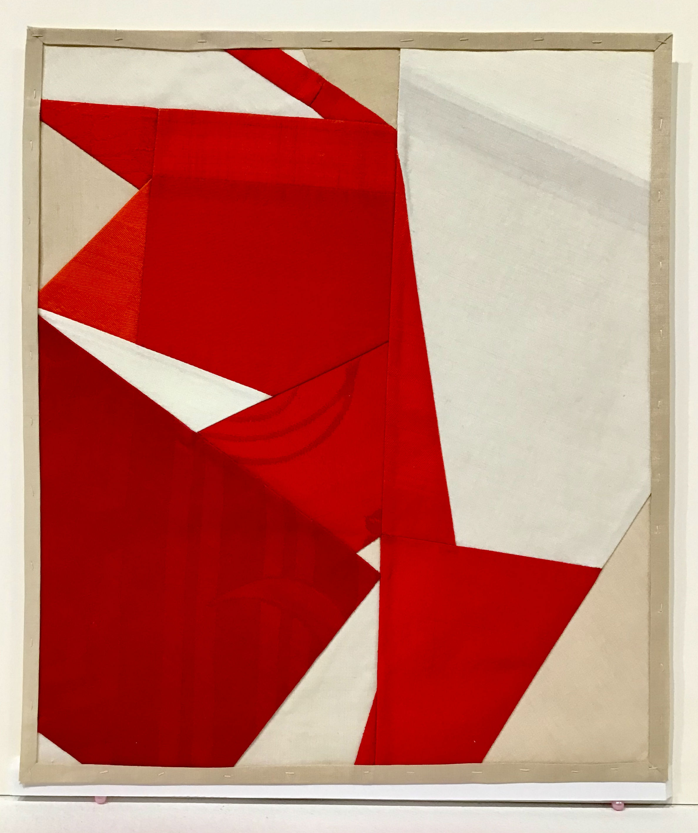 Abstract composition of pieced red and cream silk fabric arranged in angular, overlapping forms within a tan wooden frame. Sharp geometric shapes create dynamic movement across the neutral background.