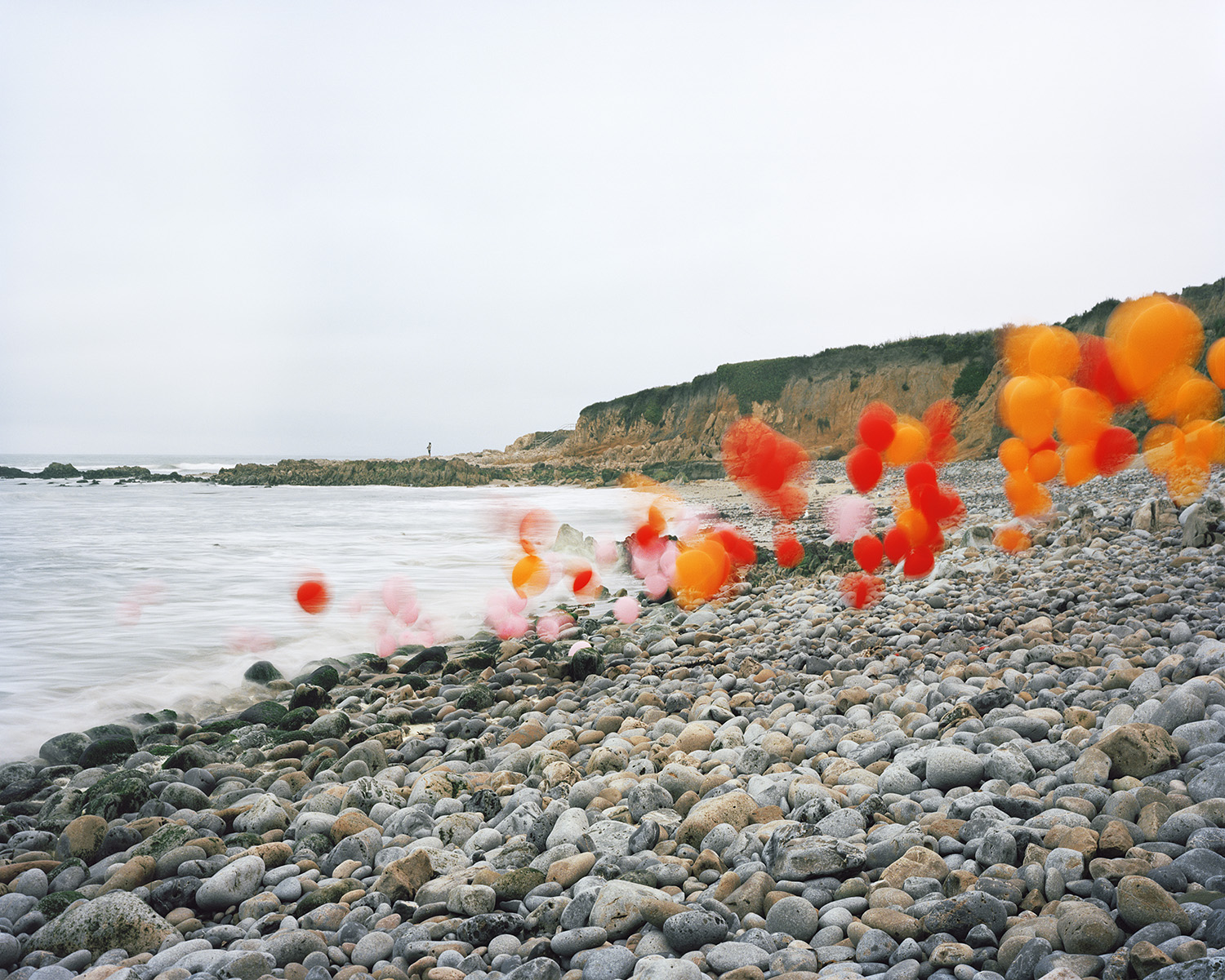Vibrant red and orange balloons float and cluster along a rocky beach, some blurred in motion, creating a striking contrast against the neutral tones of rounded stones, calm water, and distant cliffs under an overcast sky.