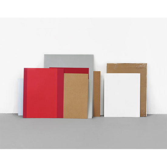 Arranged books and frames of varying heights leaning against a neutral wall, featuring red, tan, gray, and white surfaces with different textures and finishes against a gray floor.
