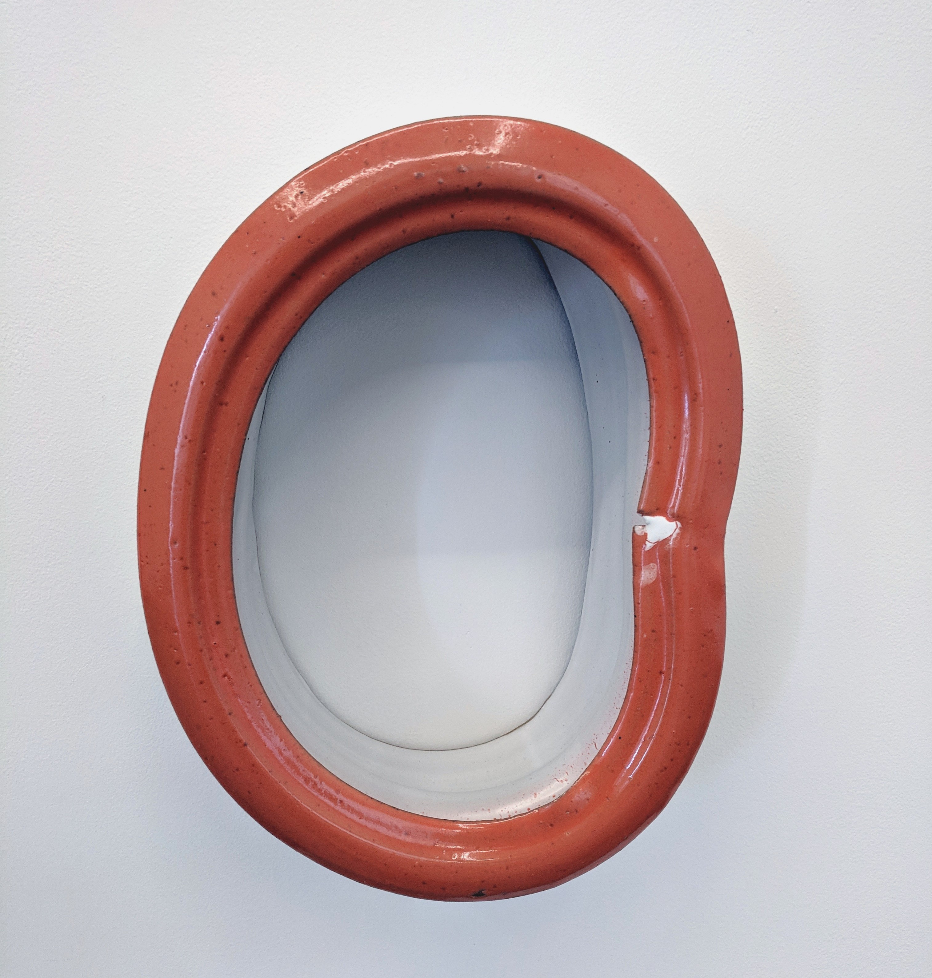 A terracotta-red ceramic form with an oval opening, featuring a smooth inner surface in contrasting gray-blue. The outer frame has a rounded, sculptural quality with visible surface texture and a small white chip on the right edge.