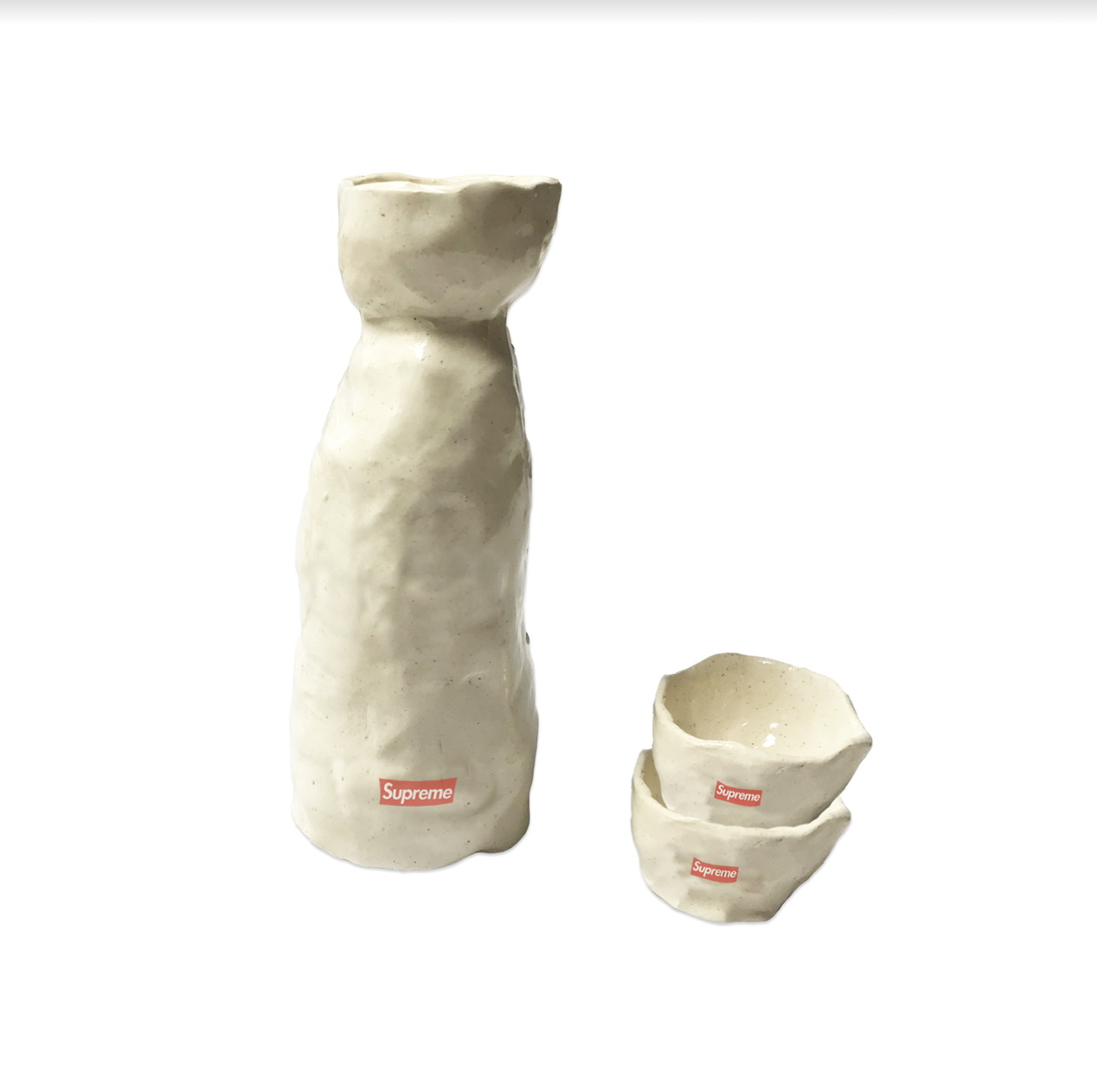 Cream-colored ceramic sake set consisting of a tall bottle with a tapered cylindrical body and flared rim, alongside two stacked small bowls with organic, slightly irregular edges. Small red rectangular logos appear on each piece against the pale, speckled glaze surface.