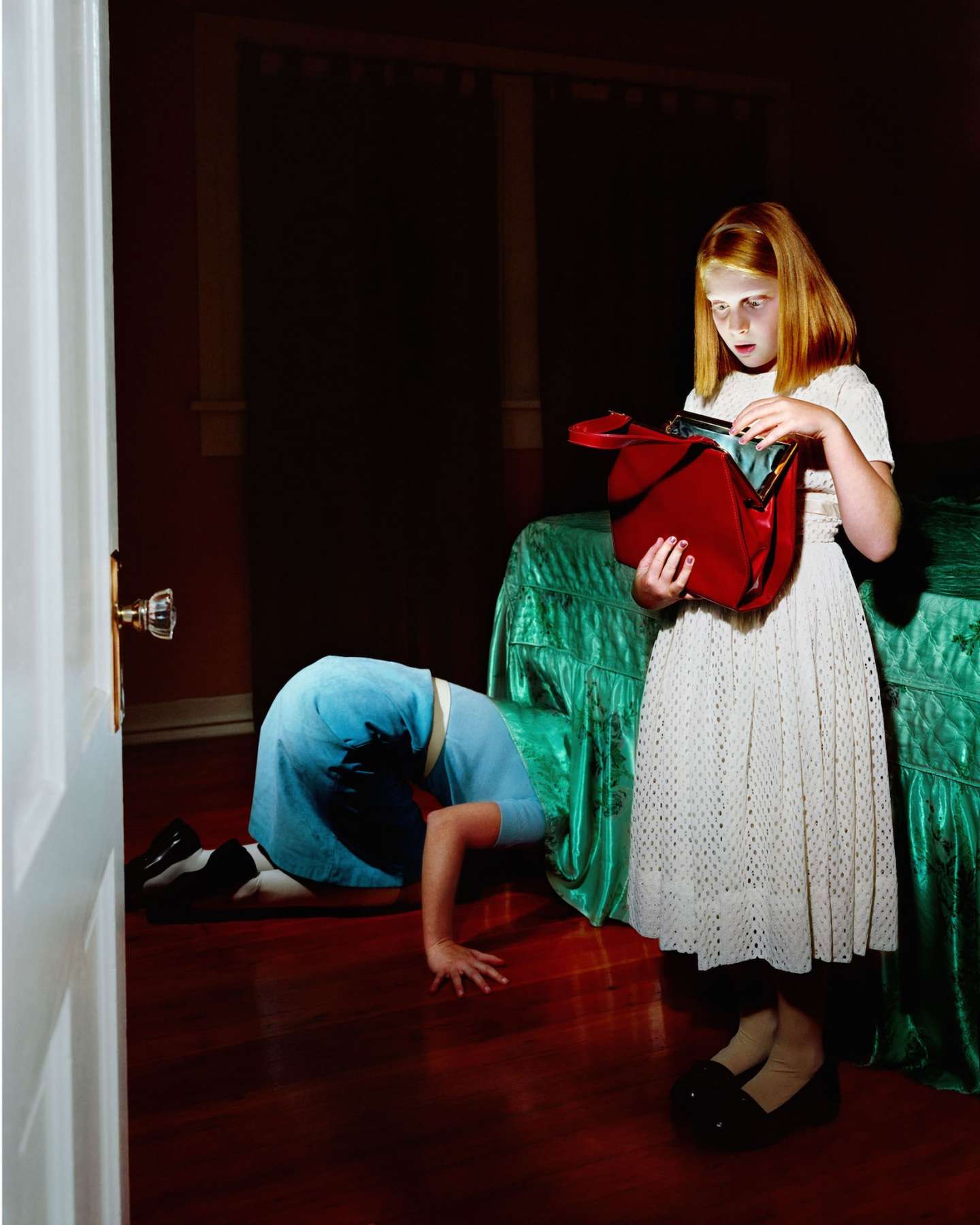 A young girl with reddish-blonde hair stands in a darkened interior space wearing a white eyelet dress, holding an open red purse. Behind her, a person in blue jeans bends forward on a green quilted fabric, their head bowed out of frame. The composition uses dramatic lighting and rich jewel tones against deep shadows.