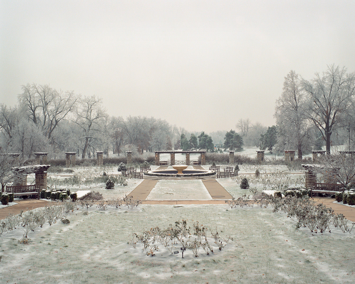 A frost-covered formal garden with a central circular fountain viewed down a symmetrical pathway. Bare trees frame the scene on both sides while brick structures and evergreens line the composition, all blanketed in ice and snow beneath an overcast sky.