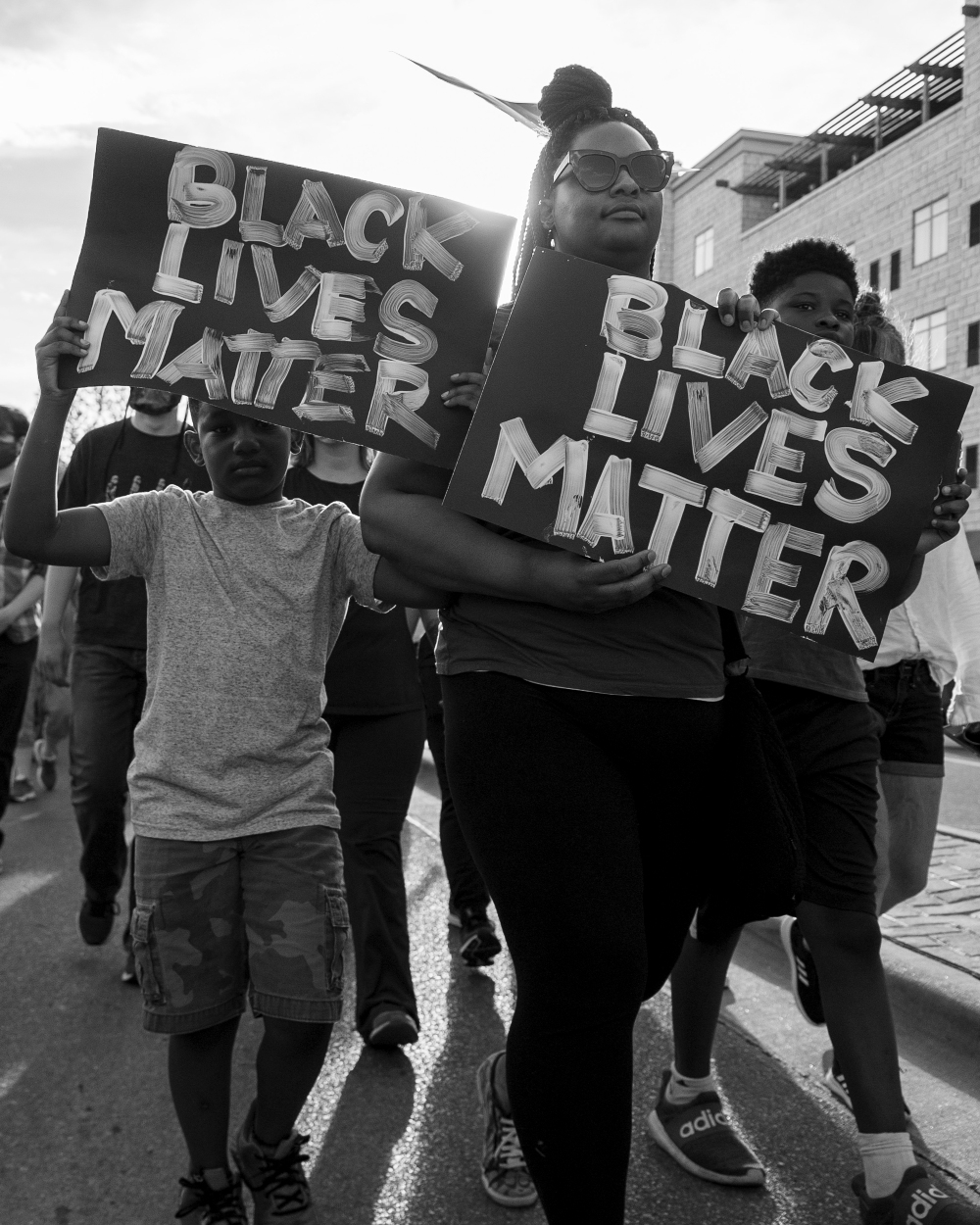 Black and white photograph of protesters marching down an urban street carrying large handmade signs reading "BLACK LIVES MATTER." The composition captures multiple participants in motion, with brick buildings visible in the background, documenting a moment of civic engagement and social activism.