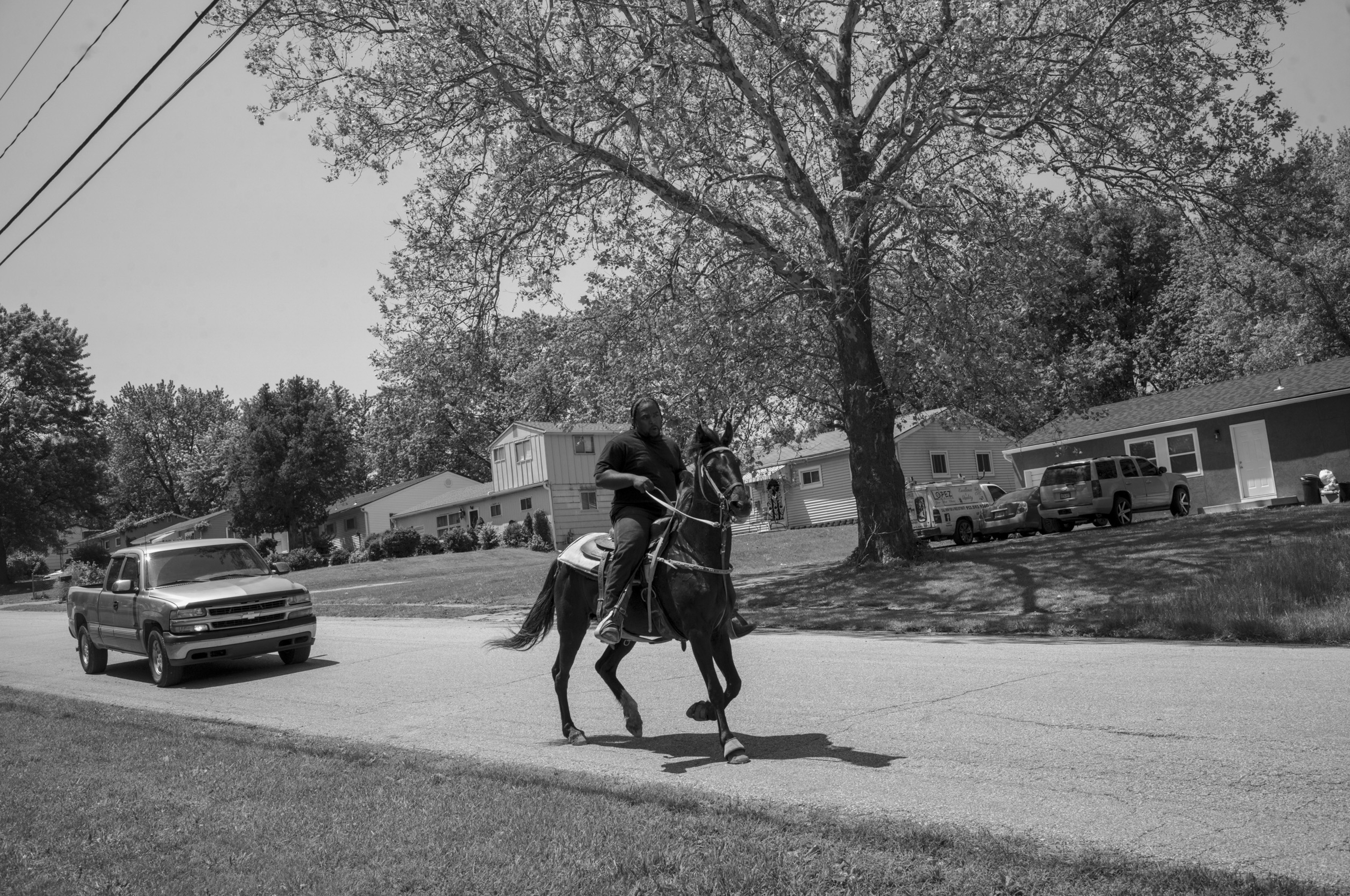 Black and white photograph of a person mounted on a dark horse trotting across a residential street lined with suburban houses and large trees, with a pickup truck visible in the foreground and utility vehicles parked nearby.