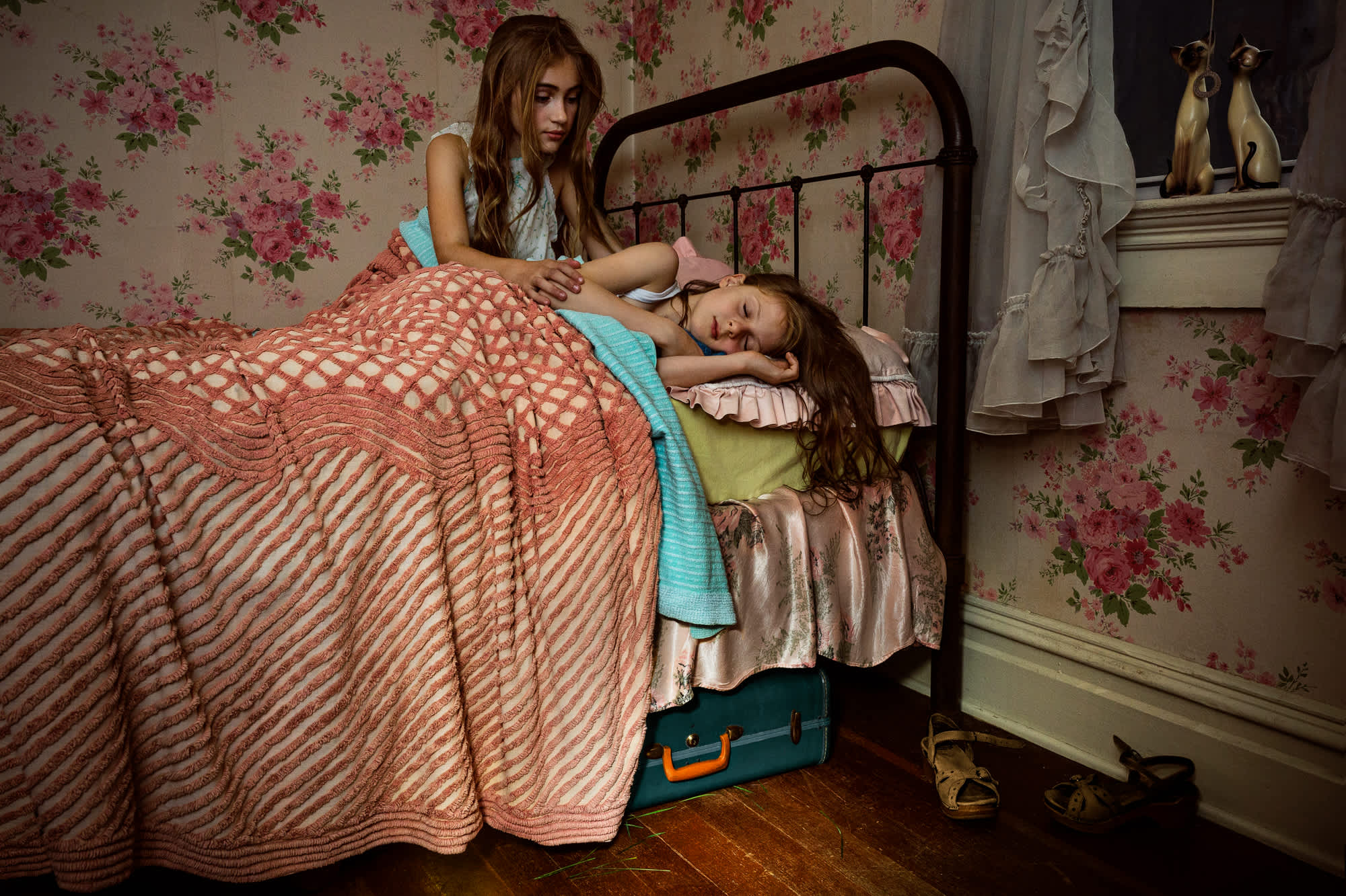 Two young girls sit on an iron bed with coral and white patterned bedding in a vintage-style bedroom. The older girl, wearing a light blue shirt, gently touches the younger girl's head as she rests on the bed. The room features floral wallpaper in pink tones, a fireplace mantel with decorative objects, and period furnishings, creating an intimate domestic scene in warm, nostalgic tones.