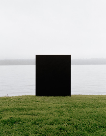A large black rectangular monolith stands centered on a grassy field, with a calm body of water and distant shoreline visible behind it under an overcast sky.