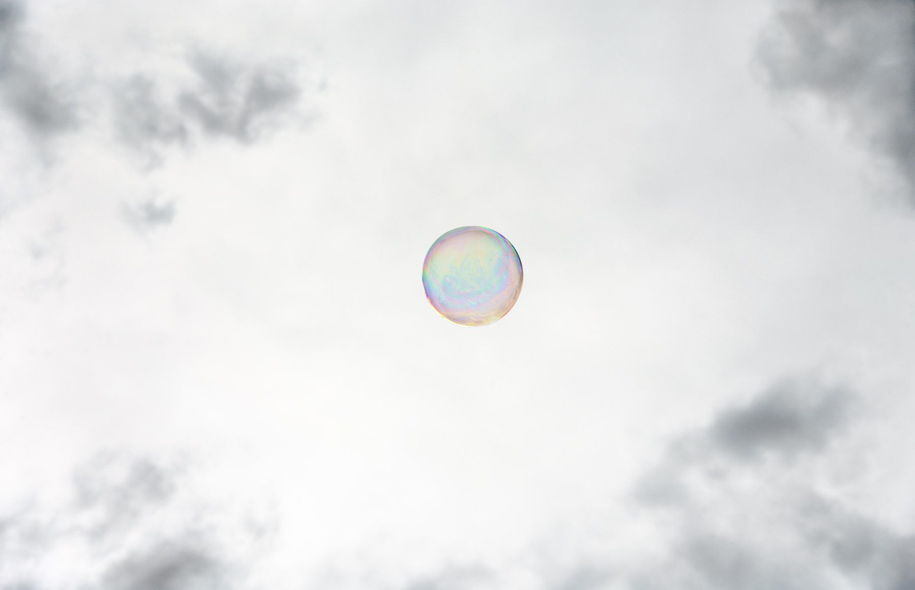 A single iridescent soap bubble floats against a soft, overcast sky with diffuse clouds visible at the edges. The bubble's translucent surface displays subtle rainbow hues of blue, pink, and yellow in its curved form.