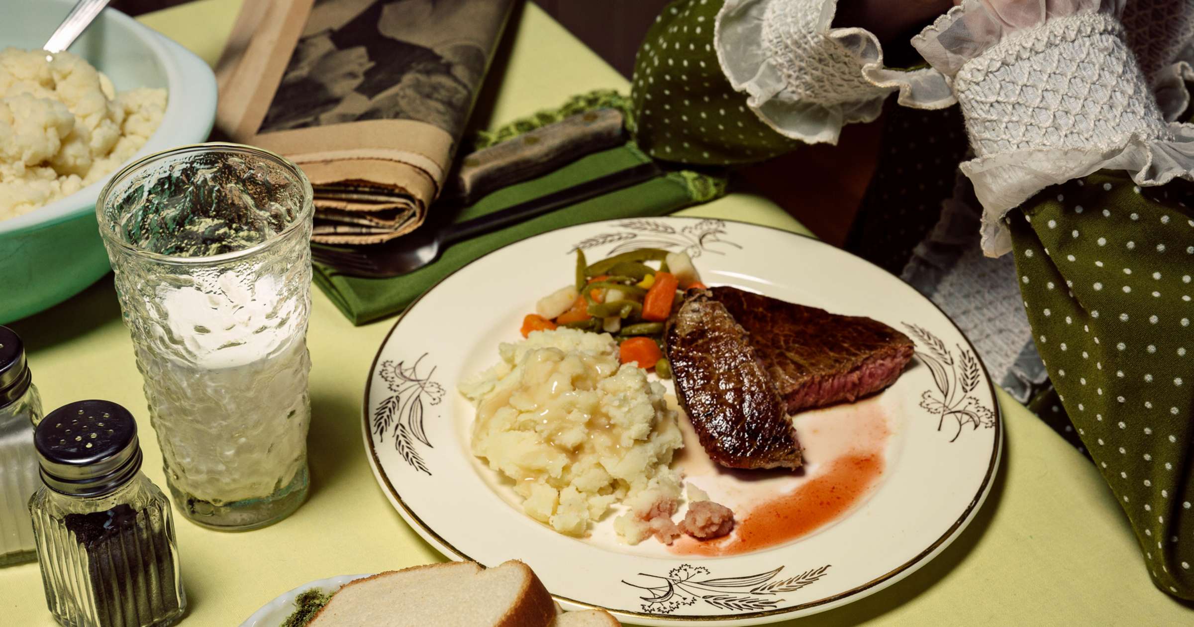 A formally plated dinner scene featuring a seared steak with mashed potatoes and roasted vegetables on a decorative white plate, accompanied by a glass of ice water, salt and pepper shakers, folded napkins, and a potted cactus with white flowers, all arranged on a pale yellow surface.