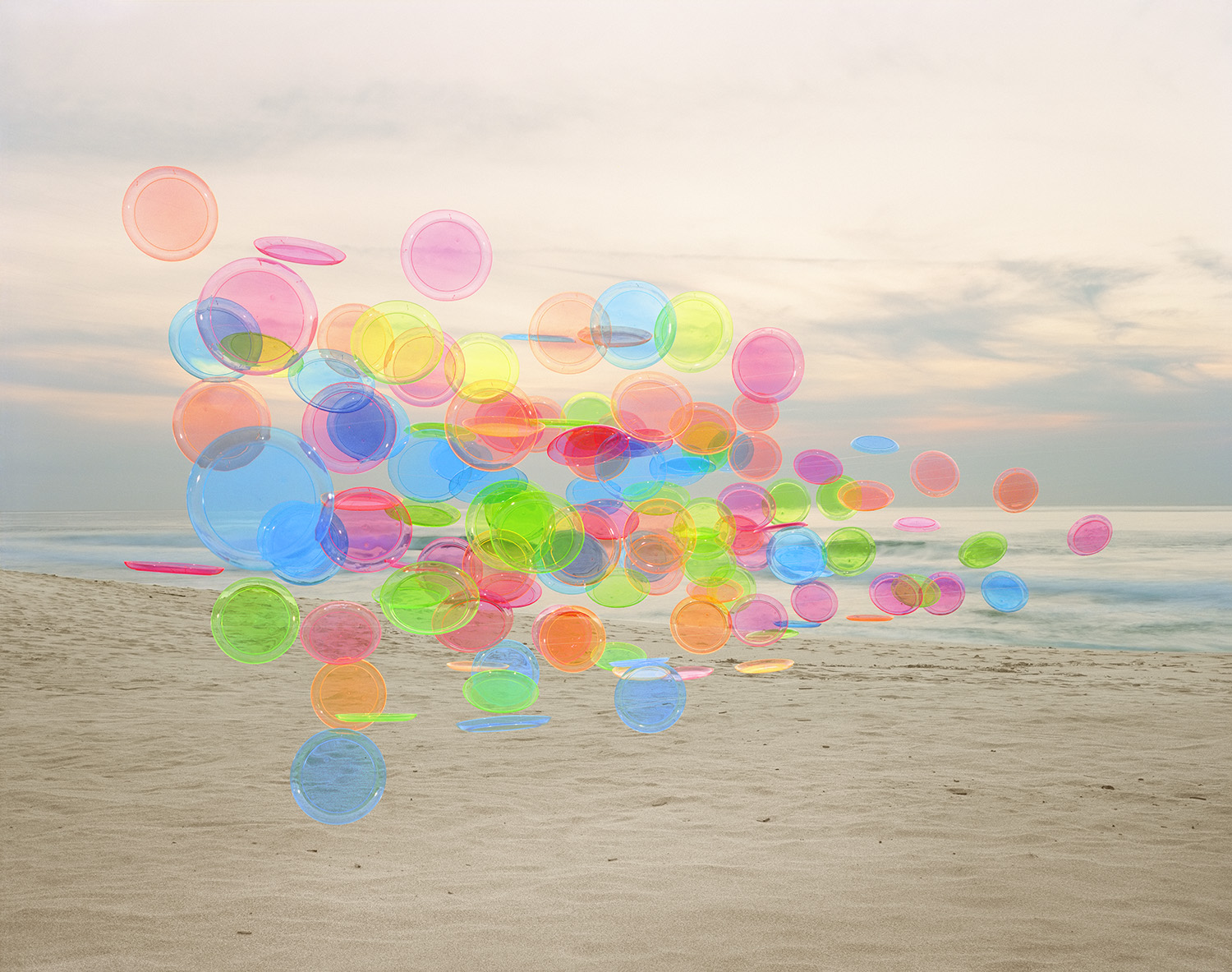 Translucent colored plastic discs and utensils in vibrant hues of blue, pink, green, yellow, and orange are arranged on a sandy beach, creating an abstract composition against a calm ocean and overcast sky.