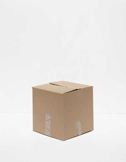 A tan cardboard shipping box photographed centrally against a neutral gray background, with visible clear packing tape on two sides and a slot cut into the top.