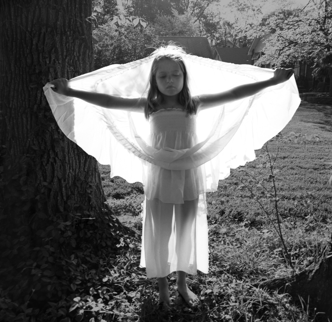 Black and white photograph of a young girl in a white dress standing in a wooded area with her arms outstretched, holding a large white fabric that billows around her like wings. Tall trees frame the composition on either side, and overgrown vegetation surrounds her on the forest ground.
