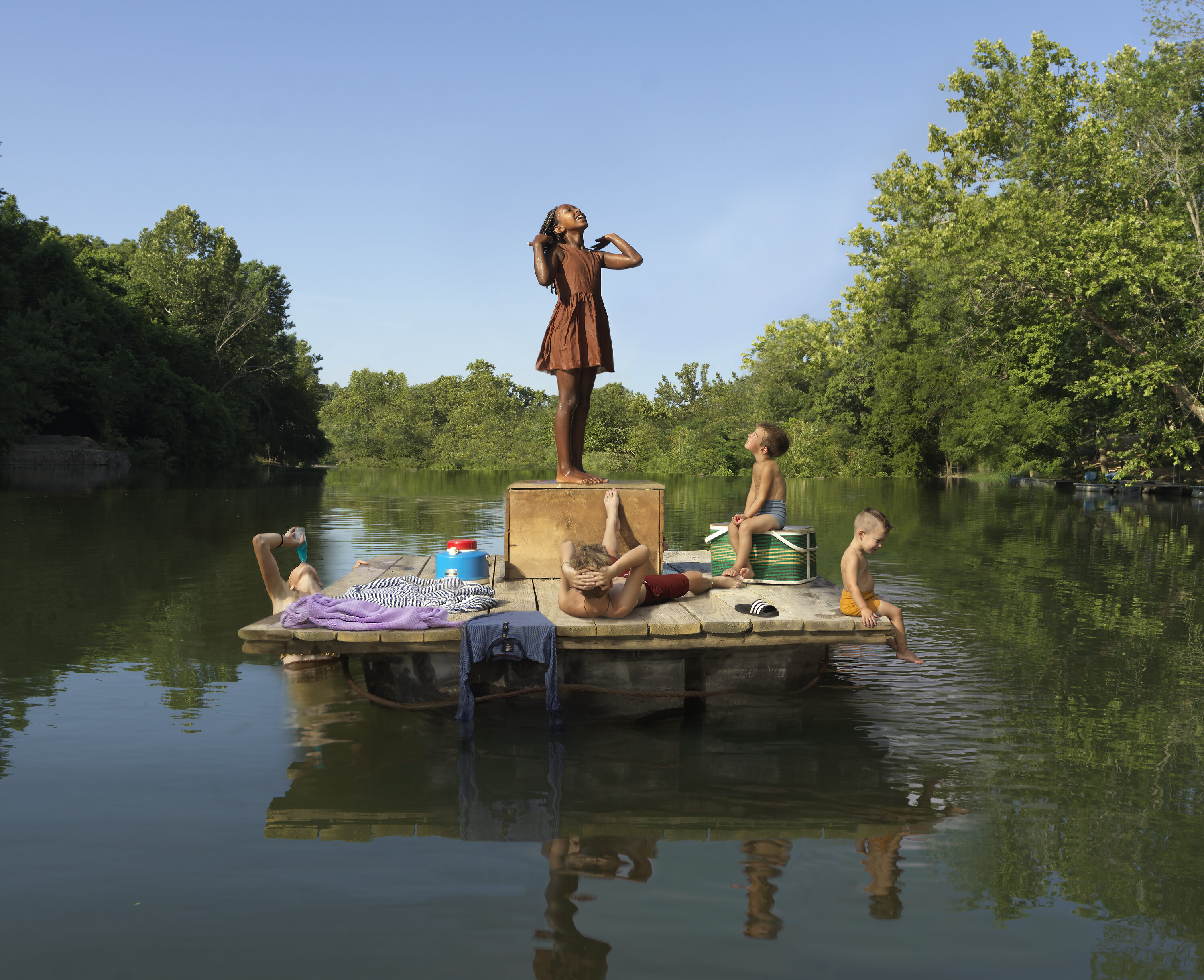 Five figures arranged on a wooden flatboat in calm water surrounded by dense green foliage. A bronze-colored statue of a standing girl dominates the center, while children in casual summer clothing lounge, sit, and play around the boat's platform. The scene captures a surreal, staged quality with warm earth tones contrasting against the cool reflective water and lush natural backdrop.
