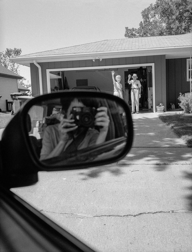 Black and white photograph taken from inside a parked car, showing a side mirror in sharp focus in the foreground reflecting the photographer's face. Beyond the mirror, a suburban house with an open garage is visible, where two figures stand waving goodbye in the driveway.