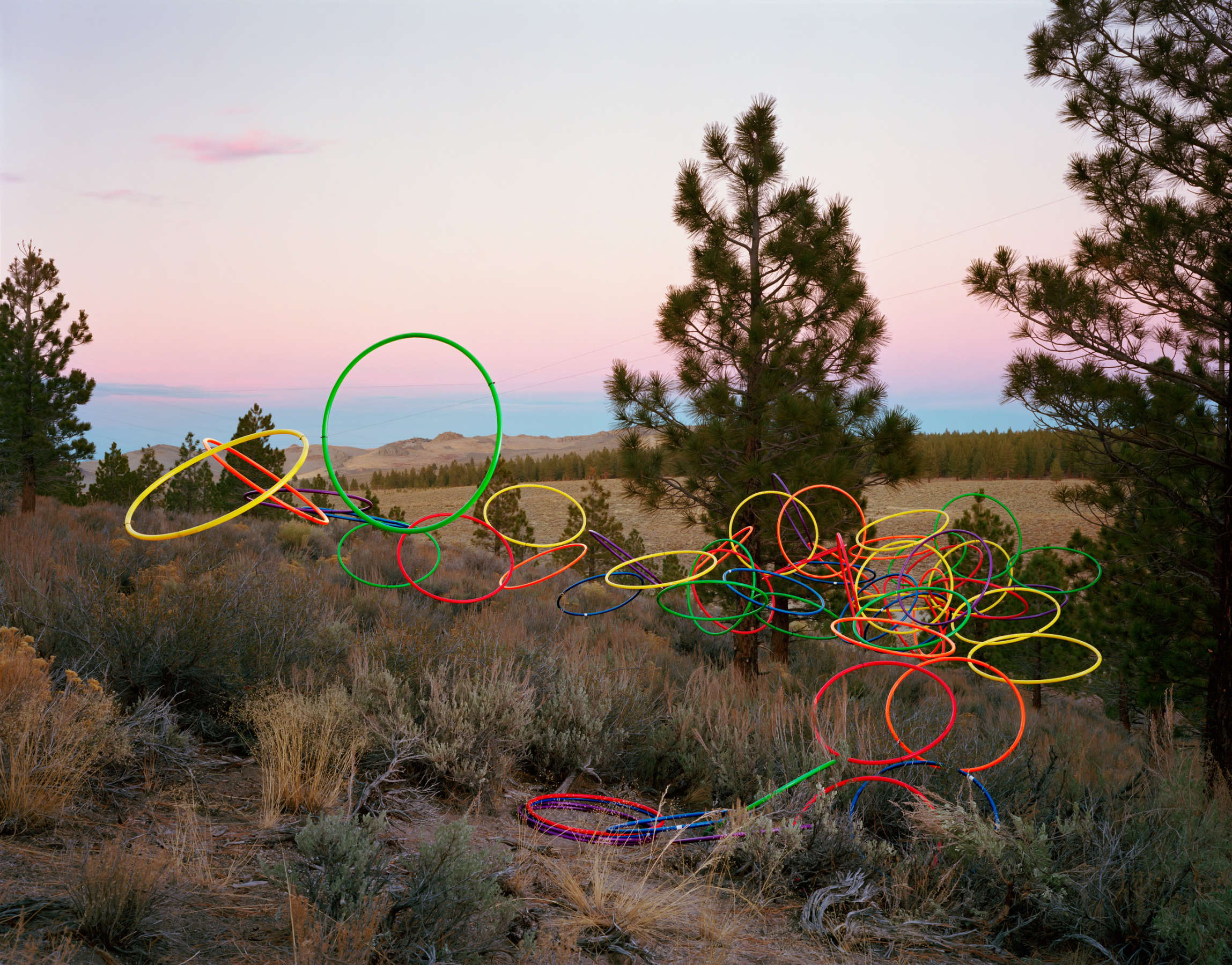 Neon-colored hula hoops in red, yellow, green, and blue arranged in an abstract composition across a desert landscape at dusk, with Joshua trees and sagebrush visible beneath the glowing sculptural forms against a twilight sky.