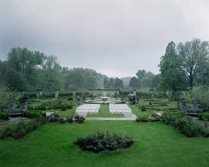 A formal garden ceremony setup with white chairs arranged in rows beneath a white tent pavilion, flanked by manicured hedges and classical stone columns. Lush green lawn and mature trees frame the scene under an overcast sky, with water visible in the distance.