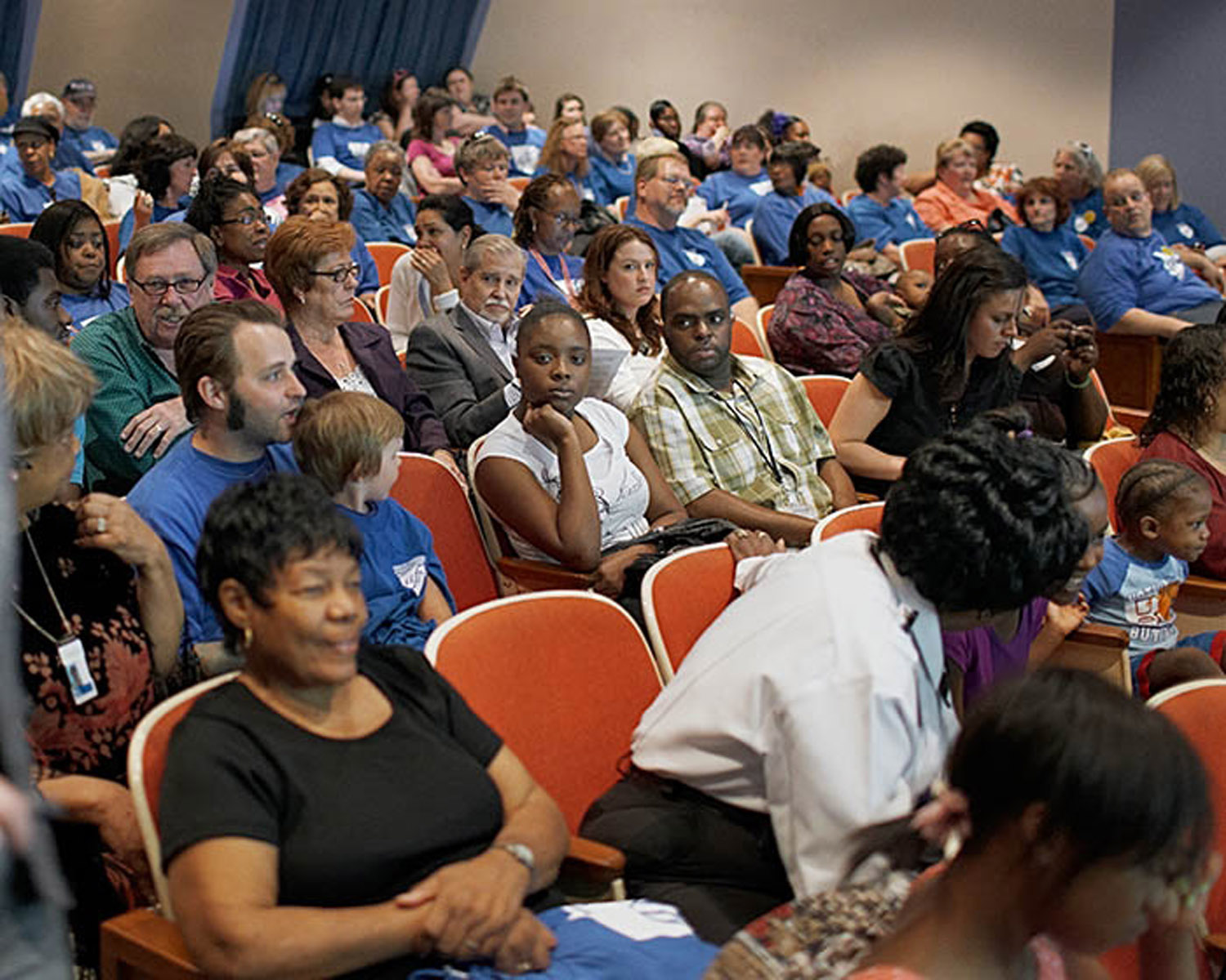 A crowded auditorium filled with diverse attendees seated in orange chairs, many wearing blue volunteer shirts. The composition captures multiple rows of people facing forward, with children, adults, and elderly individuals visible throughout the frame, creating a sense of community gathering or civic engagement.