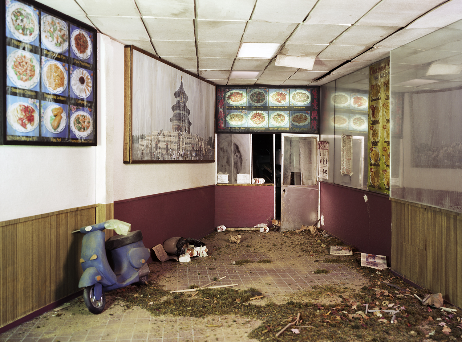 An abandoned fast-food restaurant interior filled with dirt and debris, its walls lined with illuminated menu boards displaying colorful dishes and decorative imagery of Asian architecture. A discarded blue scooter and scattered refuse rest on the earthen floor beneath a fluorescent-lit drop ceiling.