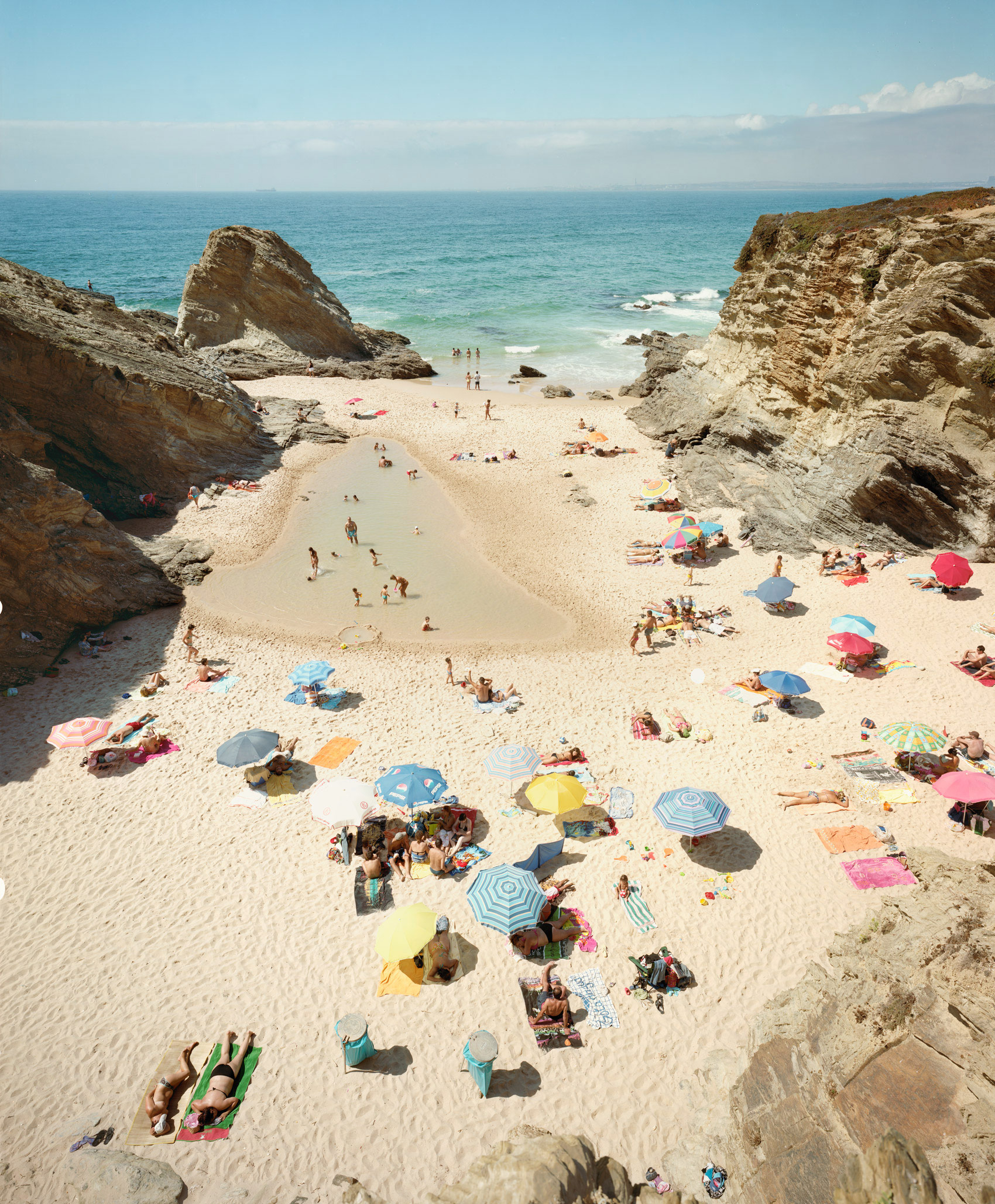 Aerial view of a crowded beach cove framed by dramatic rocky cliffs, with colorful umbrellas and sunbathers scattered across pale sand, turquoise water visible beyond.