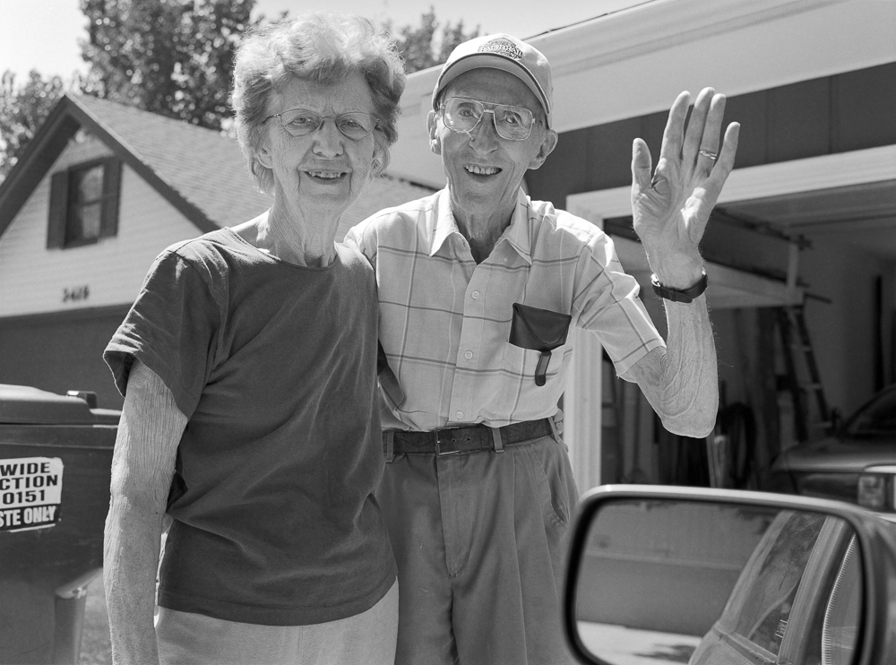 Black and white photograph of two elderly people standing together in front of an RV and residential buildings, both smiling at the camera. The man on the right wears glasses, a plaid shirt, and a cap while waving; the woman on the left wears glasses and a dark shirt. A car mirror is visible in the lower right corner.