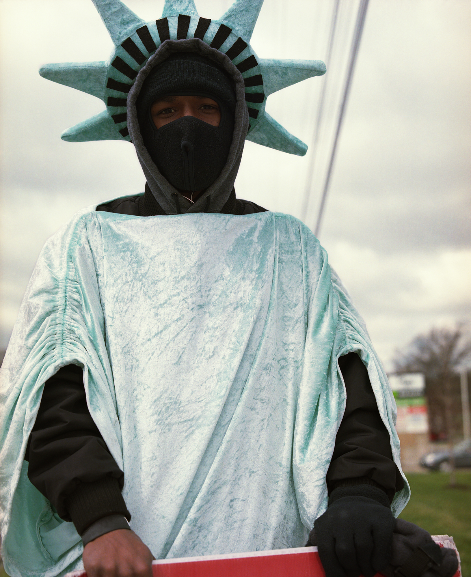 A figure wearing a custom costume of the Statue of Liberty stands facing the camera against an overcast outdoor background. The figure wears a turquoise-colored crown with radiating spikes and a draped fabric torch-bearing arm, paired with a black face mask and black hoodie, creating a contemporary reimagining of the iconic monument.