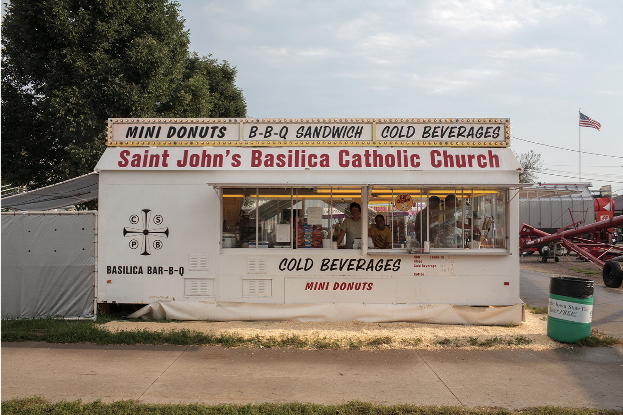 A vintage food concession stand with white exterior walls displays signage for "Saint John's Basilica Catholic Church" and "Basilica Bar-B-Q," advertising mini donuts, BBQ sandwiches, and cold beverages. The stand features large service windows and is photographed against a sky with trees and farm equipment visible in the background.