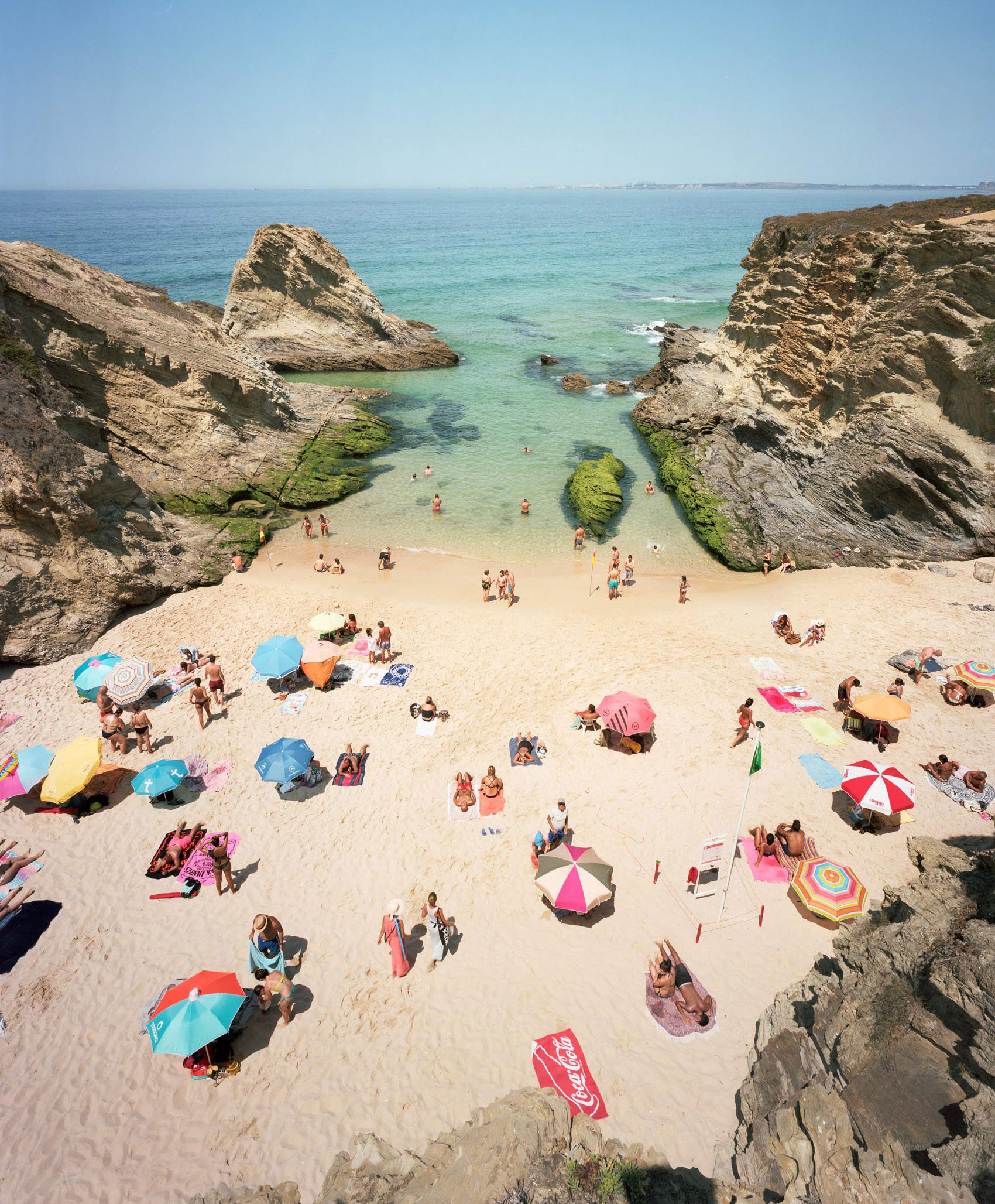 Aerial view of a crowded sandy beach nestled between dramatic limestone cliffs, with numerous colorful umbrellas and beachgoers scattered across the shore, turquoise water visible beyond with moss-covered rocks and a small island formation in the cove.